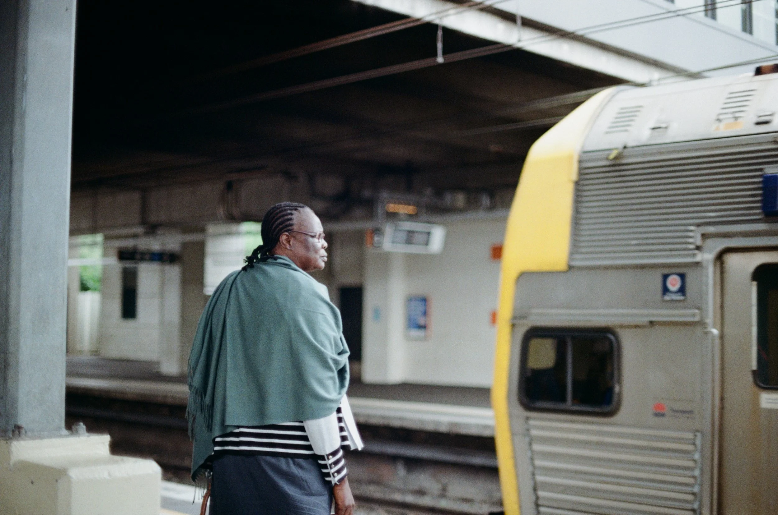 A woman with braided hair and glasses standing at a bus stop, looking to the side, with a yellow and white bus parked behind her under an overpass.