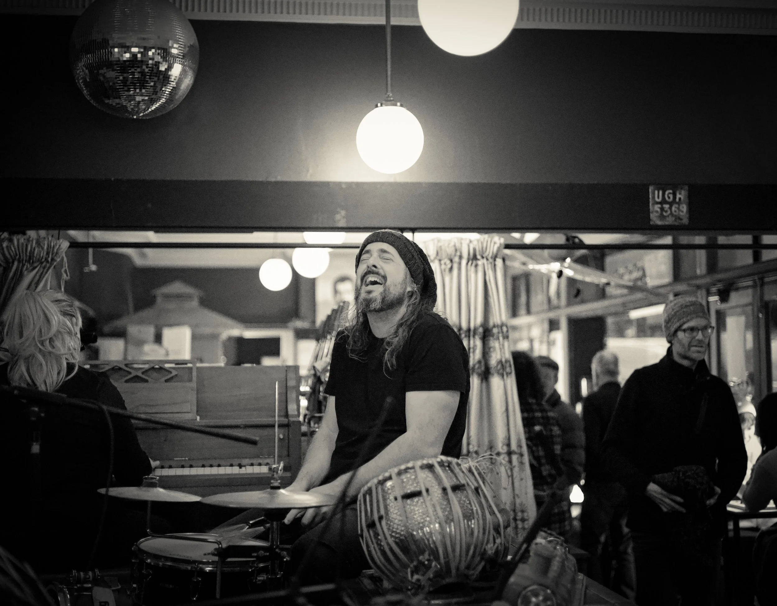 A man with a beard, long hair, and a beanie, happily playing drums at a market or indoor event, surrounded by other people and musical instruments.