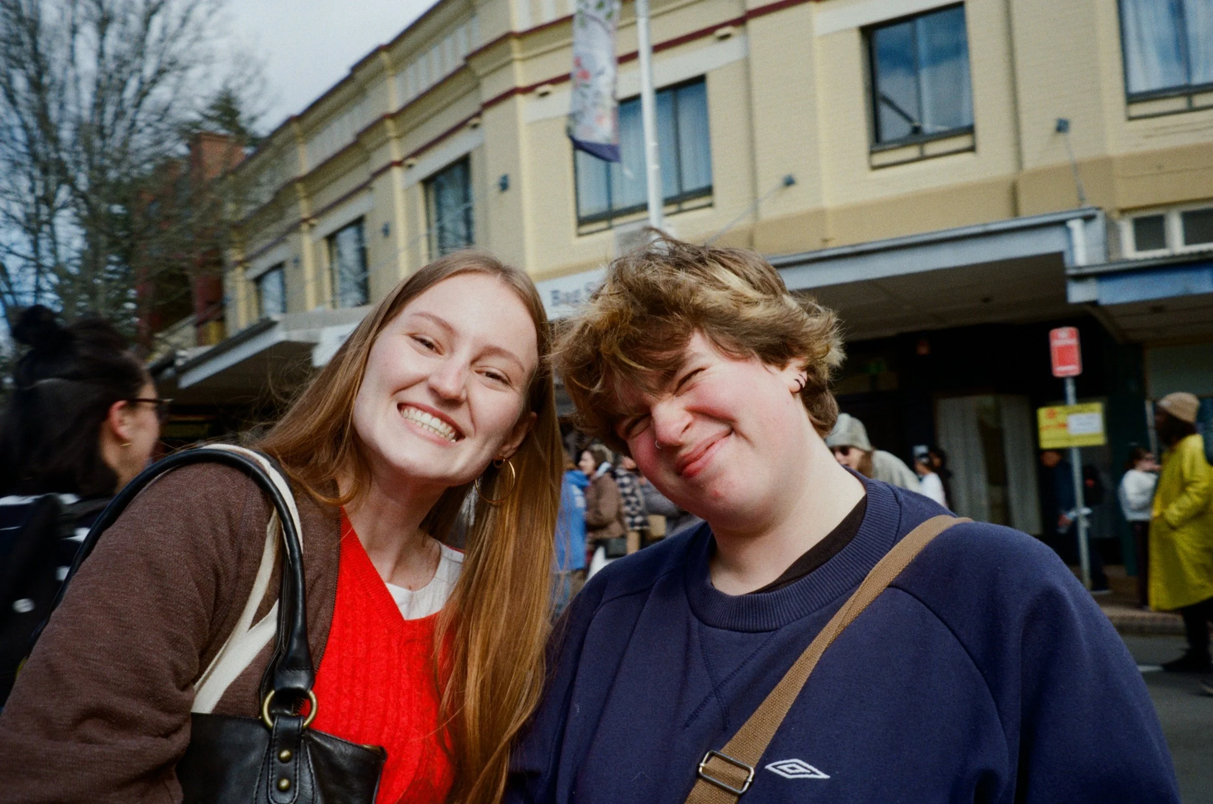 Two young people smiling and taking a selfie outdoors in a city neighborhood. The girl has long reddish hair and is wearing a brown jacket, red sweater, and hoop earrings. The boy has short, light brown wavy hair and is wearing a navy blue sweater wi