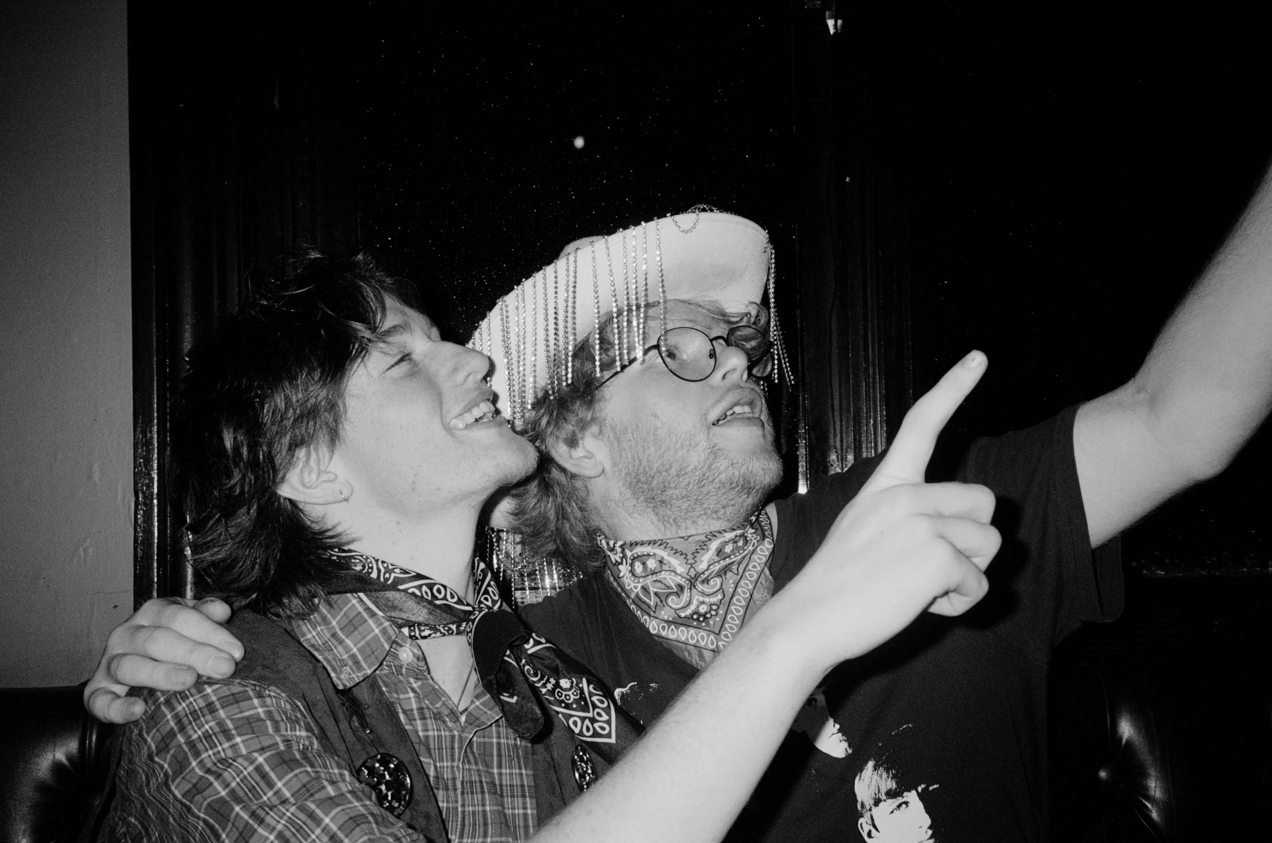 Two young men taking a selfie in a dimly lit setting. One is wearing a hat with dangling chains, glasses, and a bandana around his neck, while the other is smiling and has his arm around him. They are both smiling.