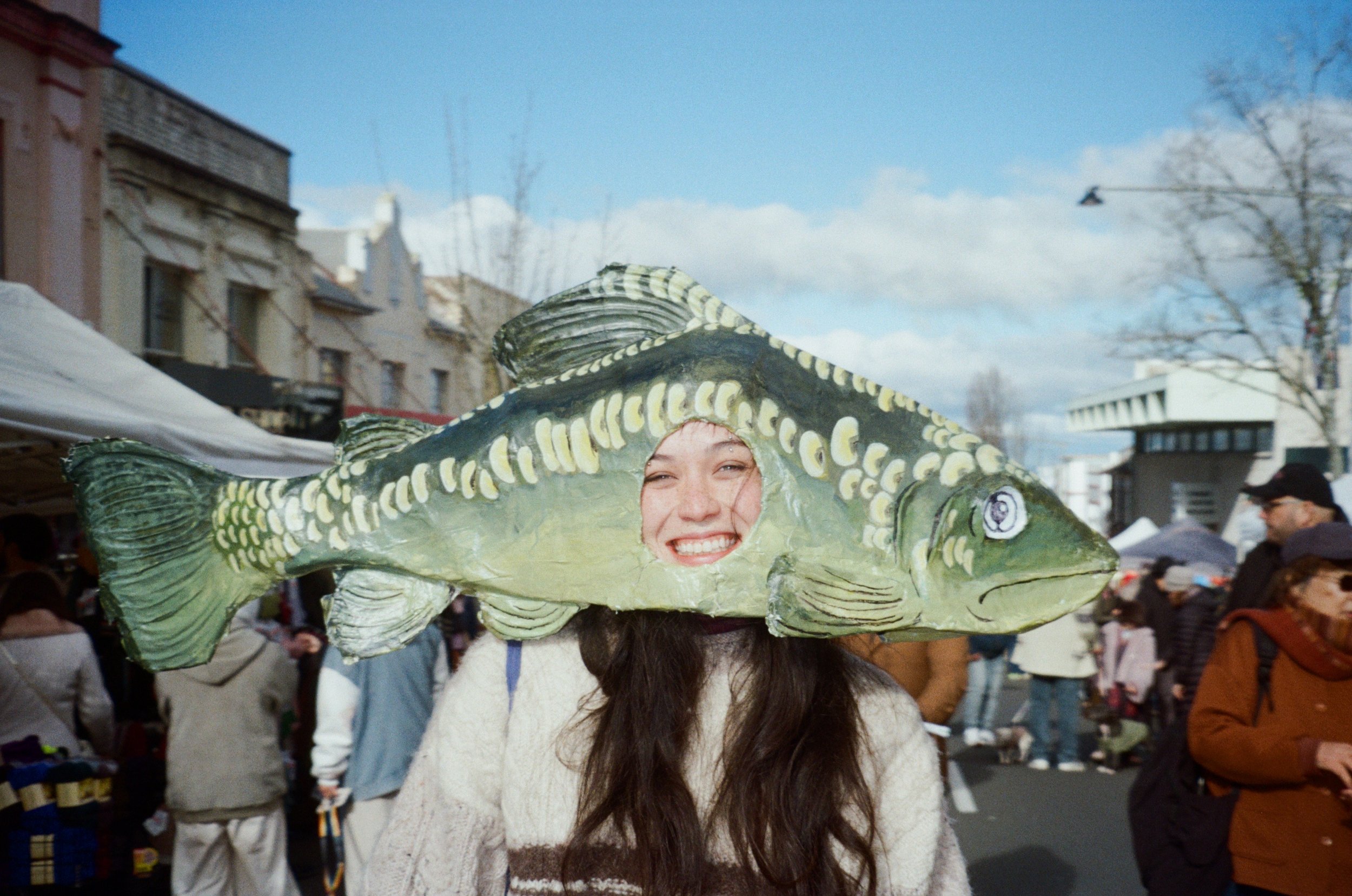 A woman wearing a large green paper mache fish headpiece at a winter festival among many people and on a crowded street