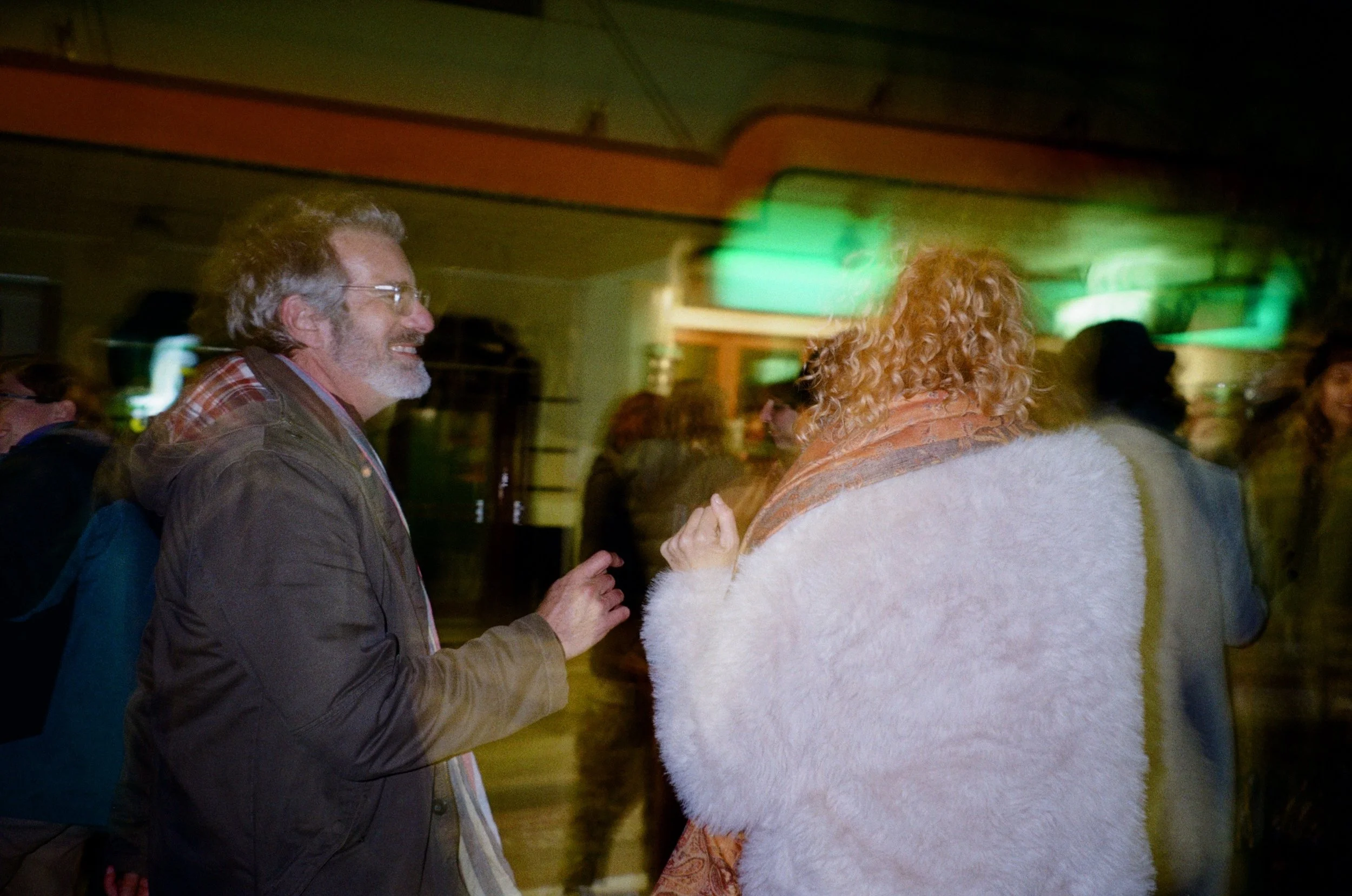 A man with glasses and a woman with curly hair and a fur coat are dancing together at an indoor event, with other people visible in the background.