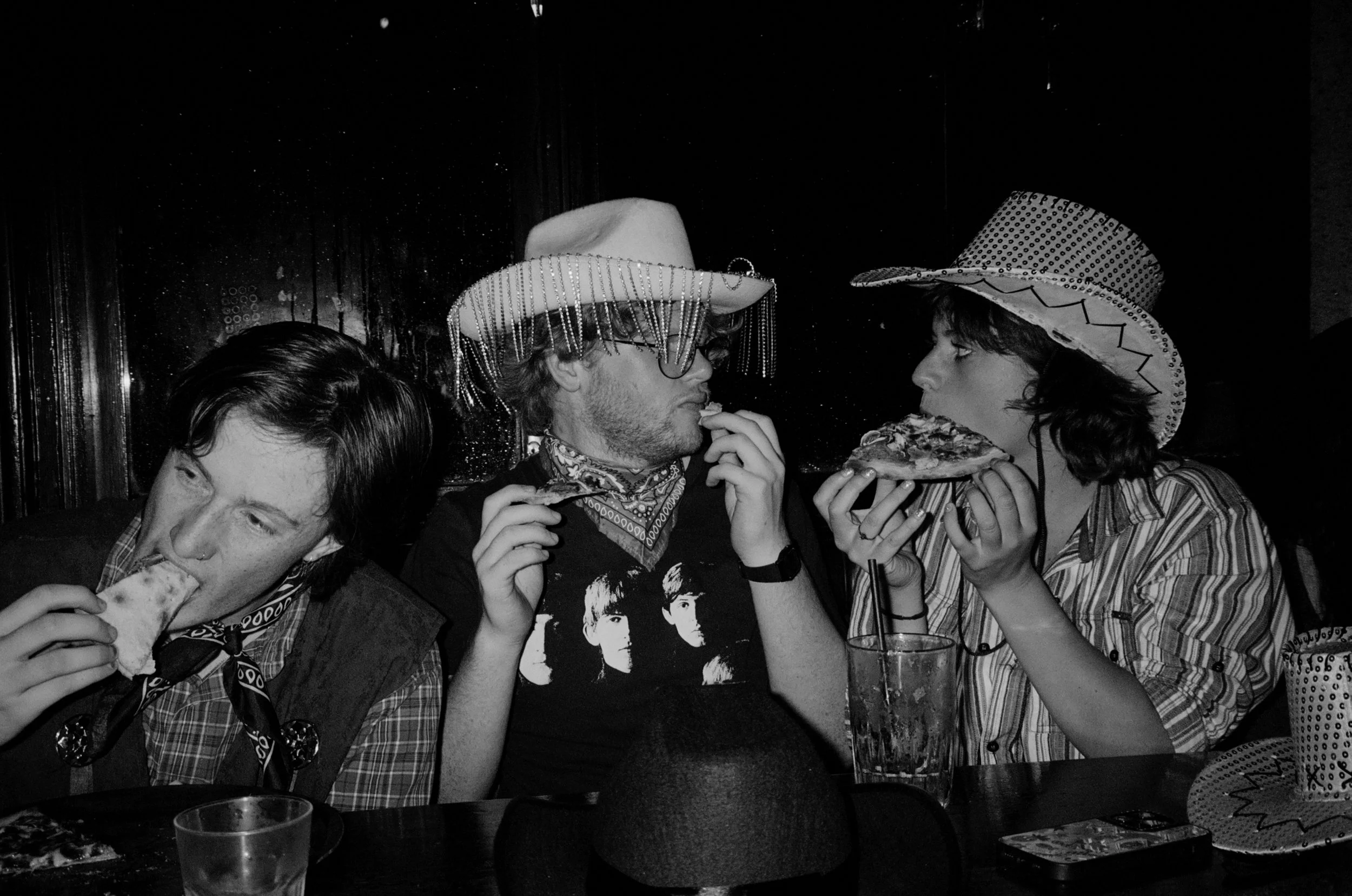 3 people conversing with one another in cowboy and western attire, while eating pizza and drinking alcohol at a table in a pub/bar setting.