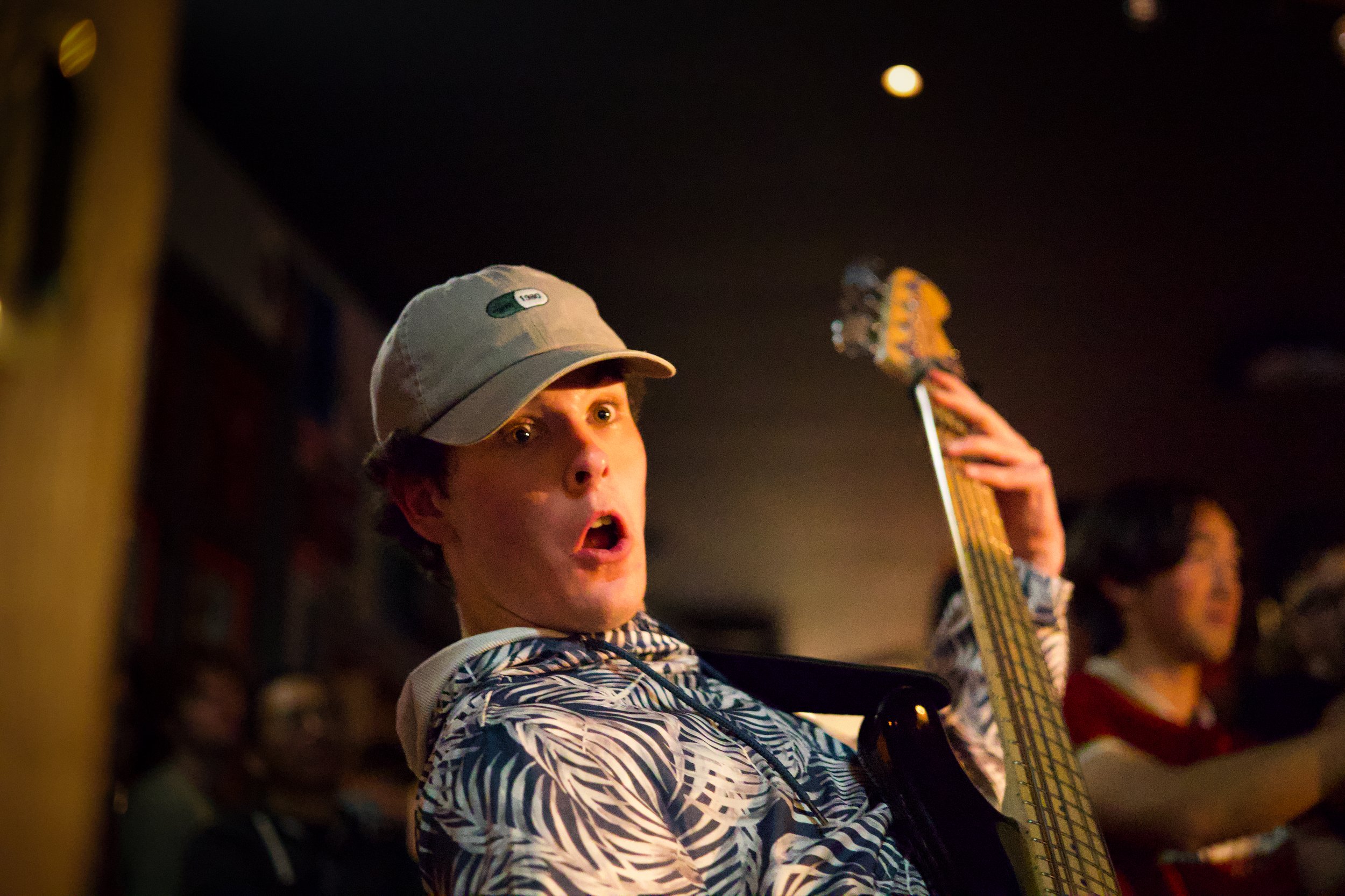 A young man wearing a grey baseball cap and patterned windbreaker jacket appears surprised or excited, holding a bass guitar, in a dimly lit setting with other people in the background.