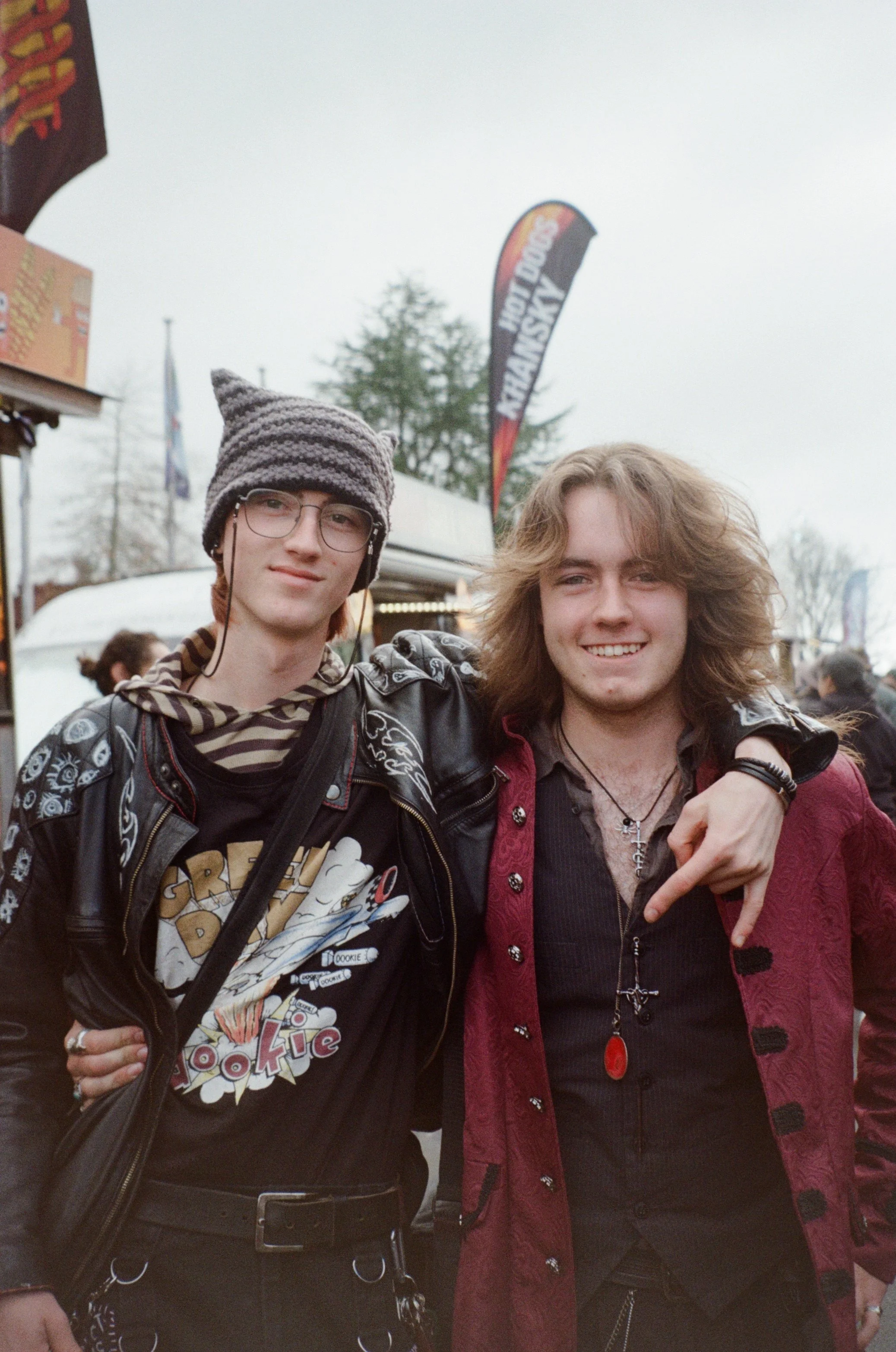 Two young men standing close together outdoors at an event, smiling at the camera. One is wearing a striped beanie, glasses, a leather jacket, and a graphic T-shirt; the other has long curly hair, a red quilted jacket, and a necklace.