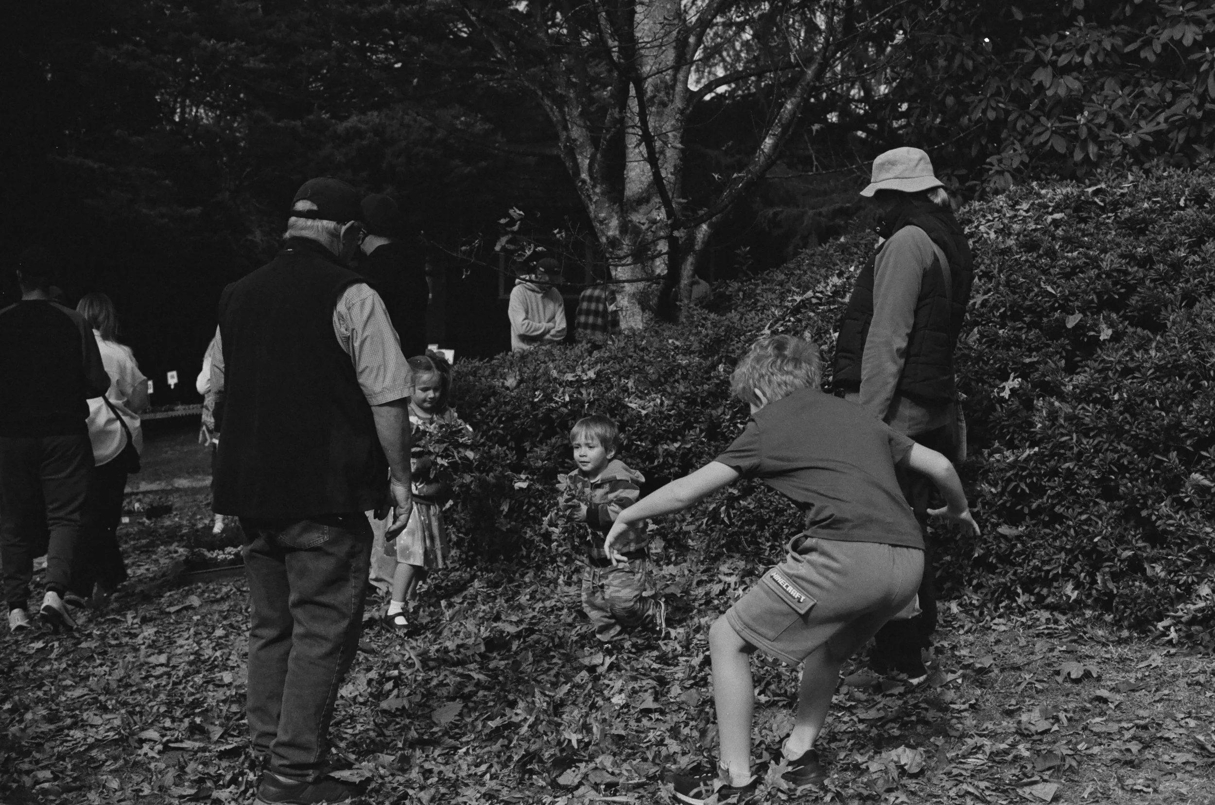 A group of people, including children, gathered outdoors under trees, appearing to be involved in an activity or discussion. The scene is in black and white.