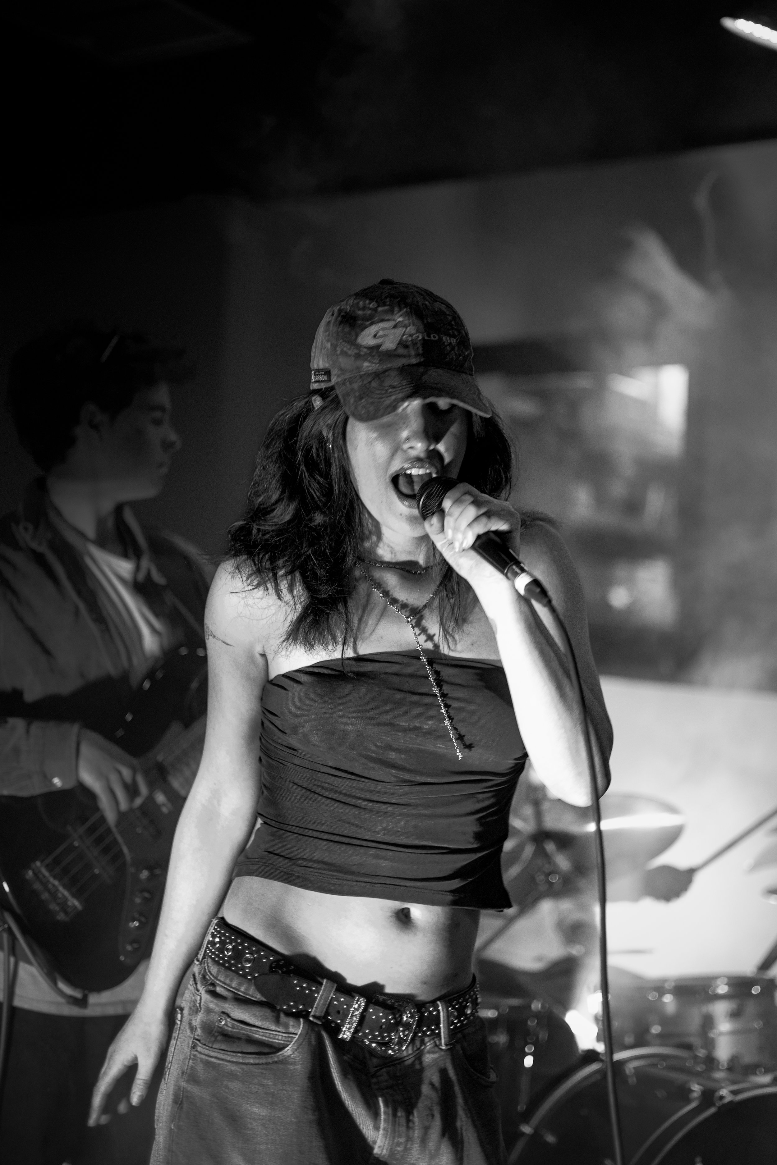 A woman singing into a microphone on stage, wearing a cap and a strapless top, with a person playing guitar in the background.
