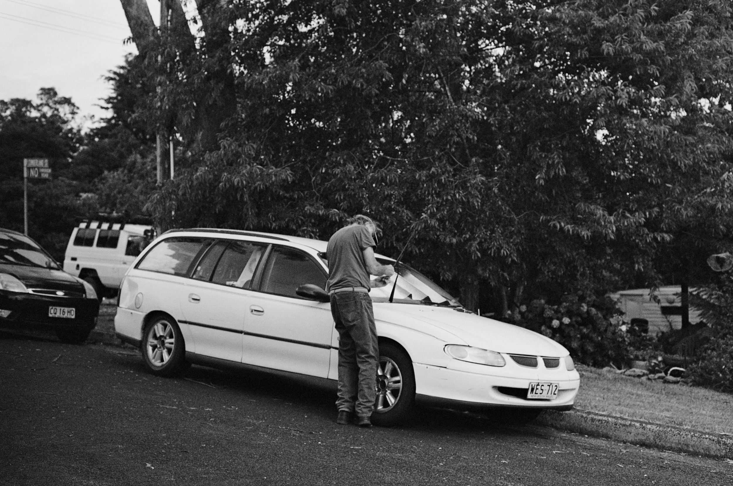 A man is checking the windshield wipers of a white car parked in suburban street during daytime, with multiple vehicles and trees in the background.