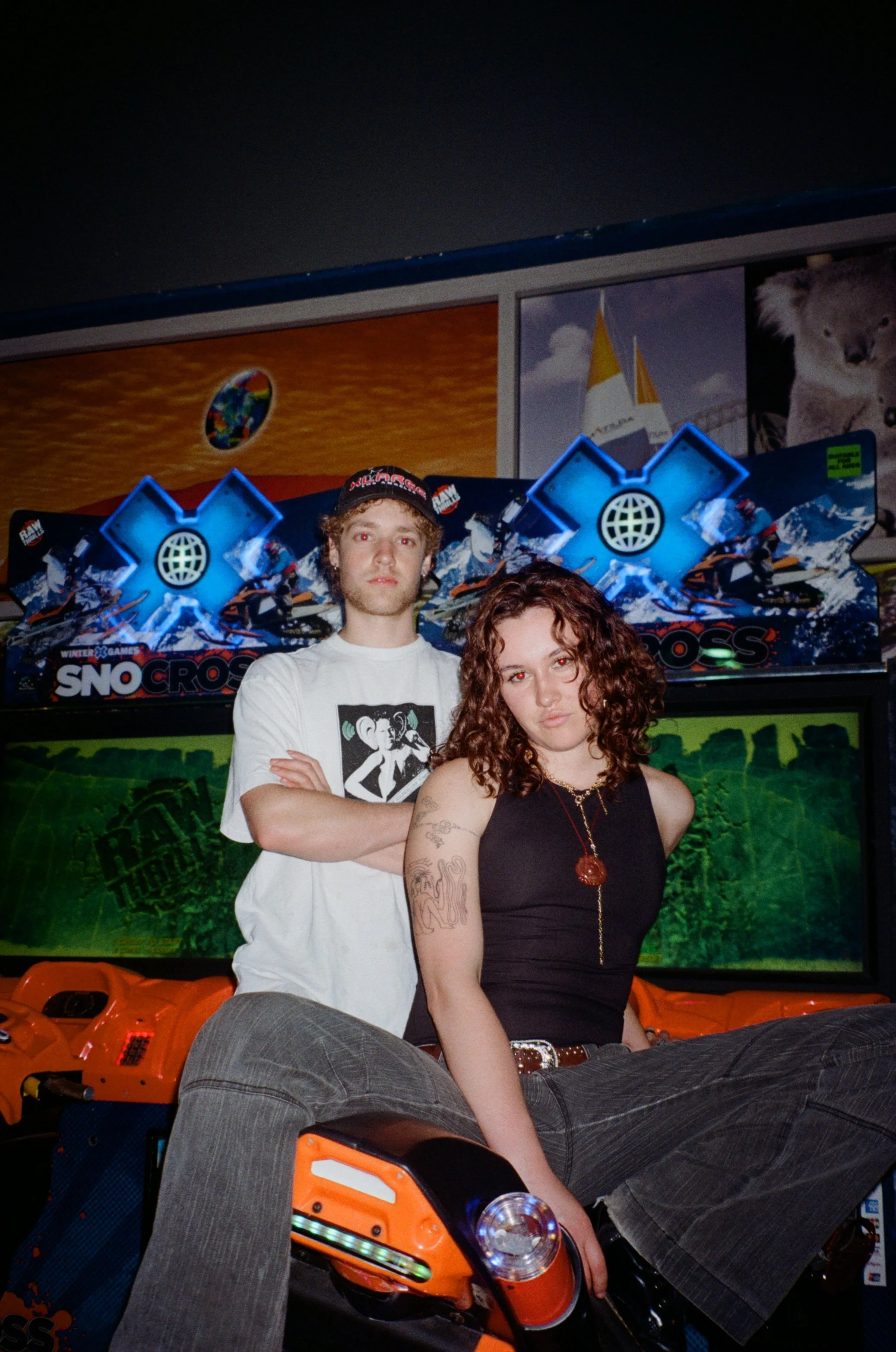 Two young adults posing in front of a colourful arcade game, with one person sitting on the arcade's motorcycle arcade game and the other standing behind with arms crossed, in an indoor arcade/cinema setting.
