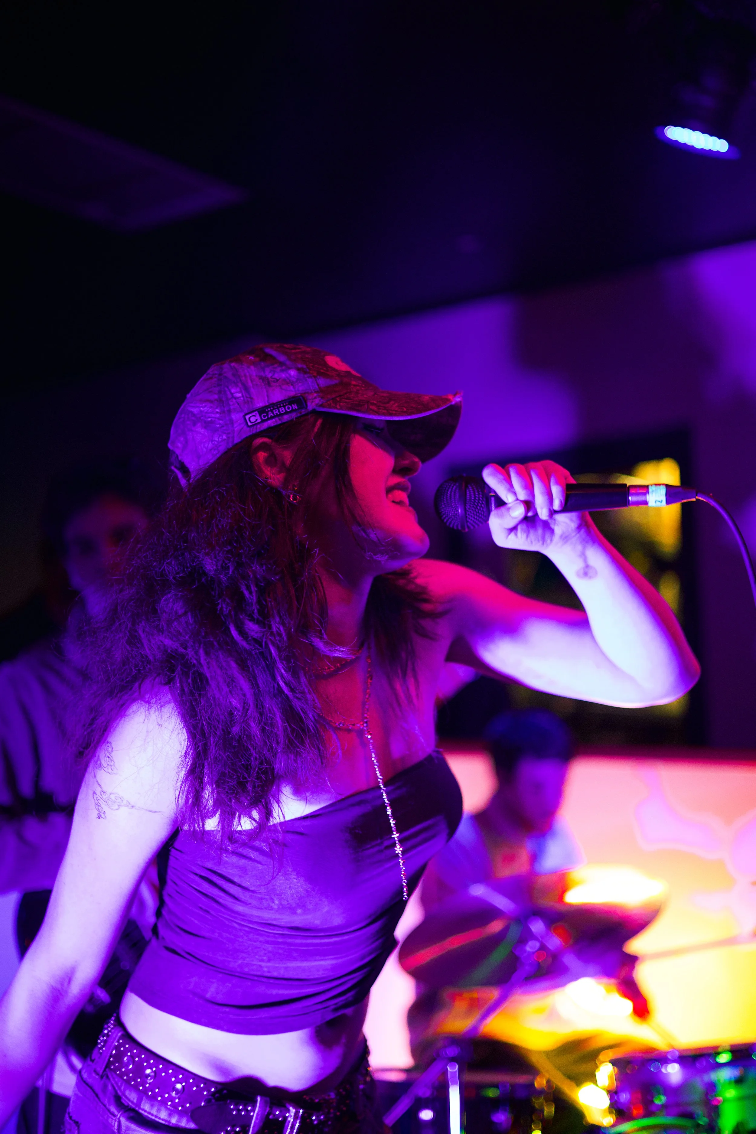 A woman singing into a microphone at a nightclub or music venue with purple and blue lighting, wearing a camouflage cap, a striped tube top, and a studded belt, with drums in the background.