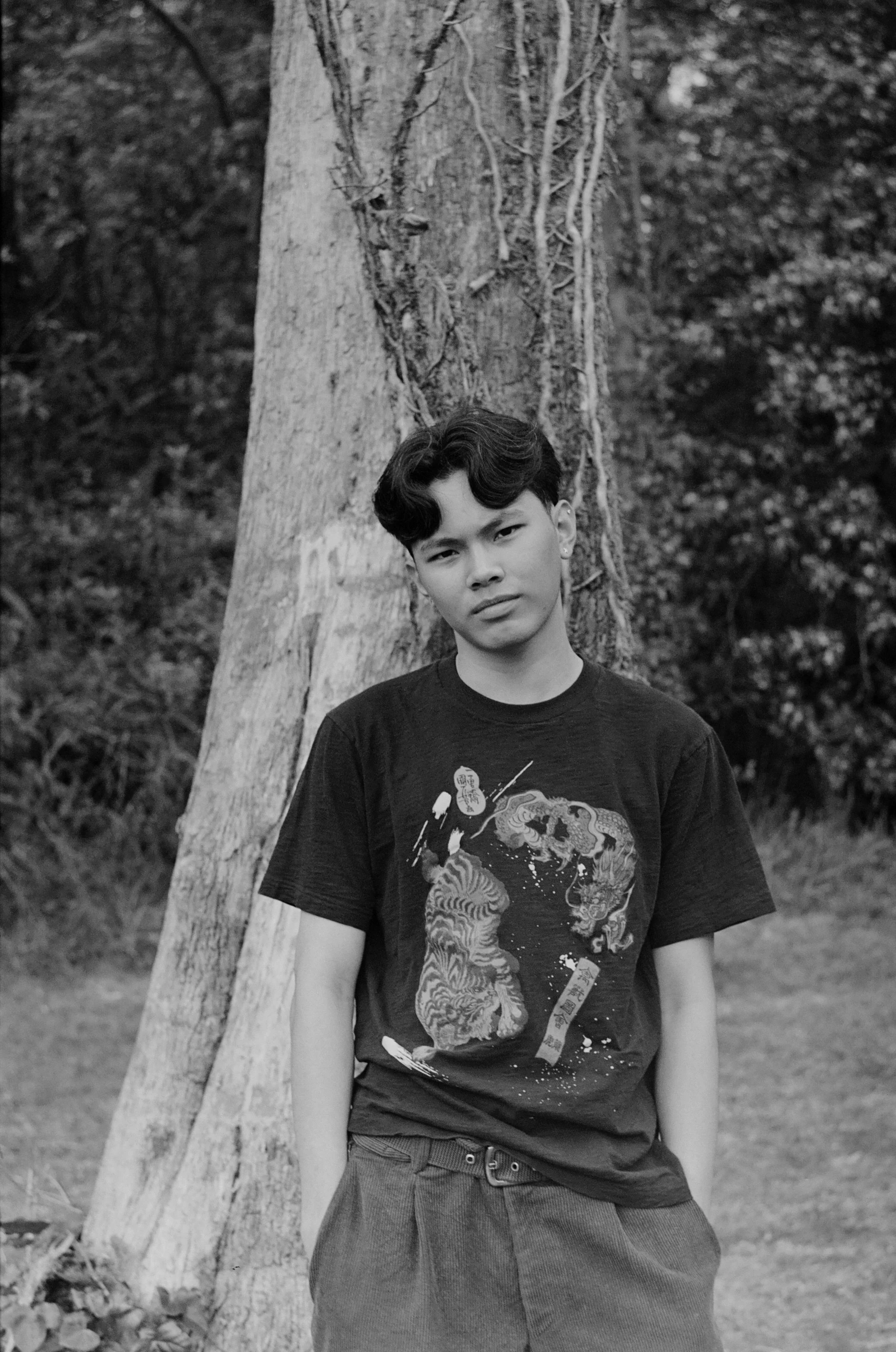 A young man with wavy hair standing outdoors with a large tree in the background. He is wearing a graphic t-shirt and loose-fitting pants, looking at the camera with a slight frown.