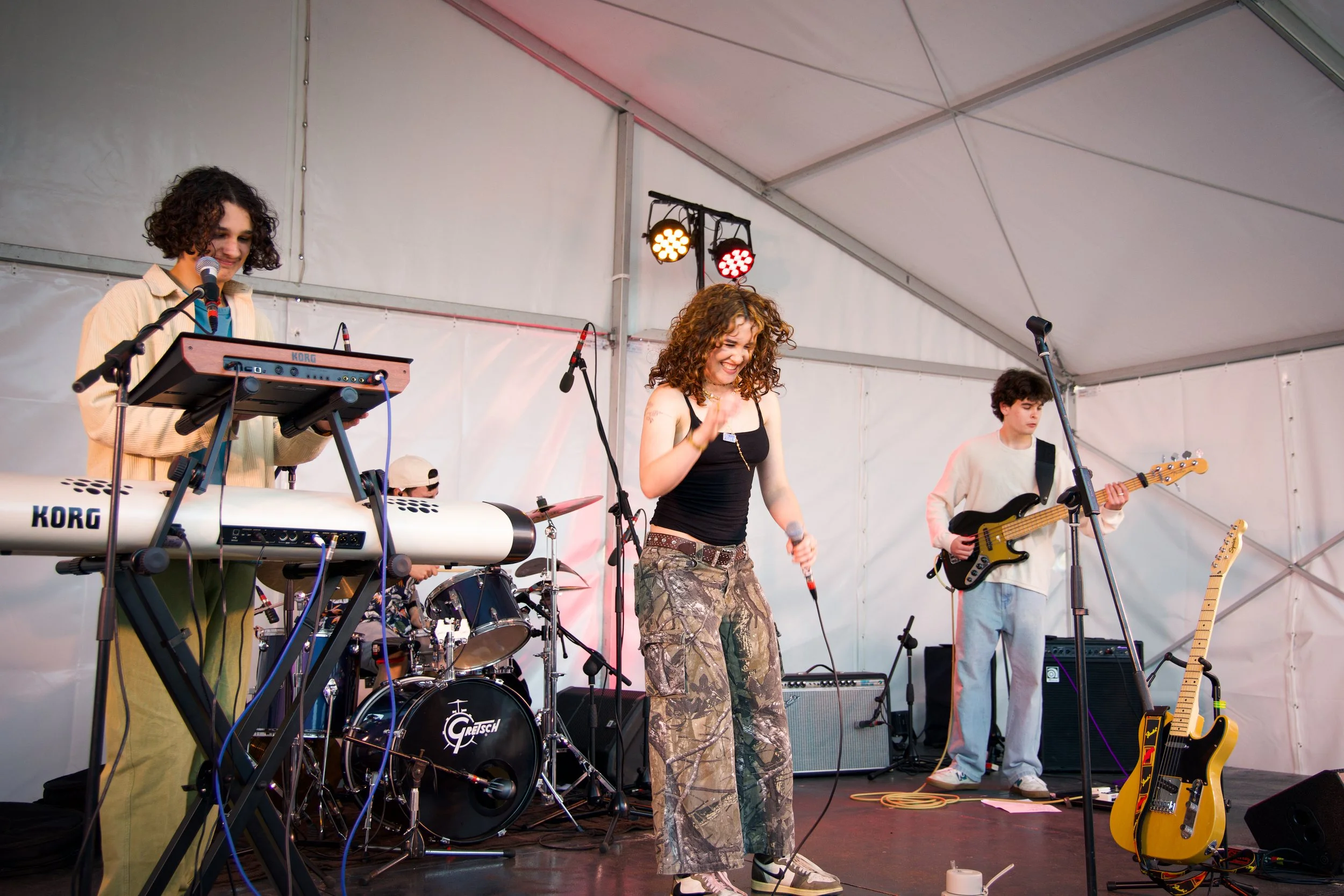 A band of five members performing on a stage inside a white tent. One member is at a keyboard, another at drums, a woman in the center is singing into a microphone, a guitarist, and another member is partially visible. The stage has various musical e