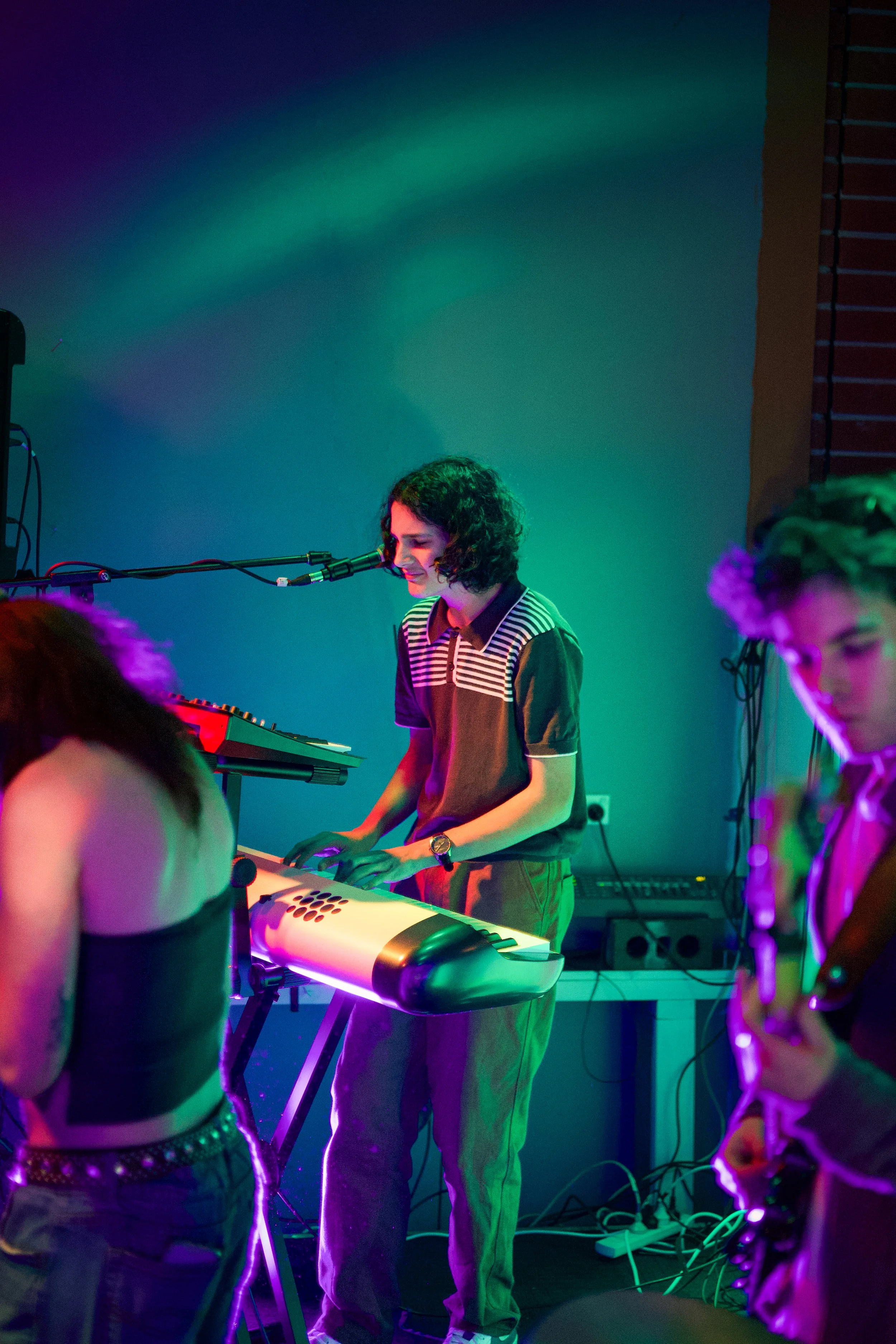 A woman with short, curly hair playing a keyboard at a music event with colorful lighting, with other people partially visible around her.