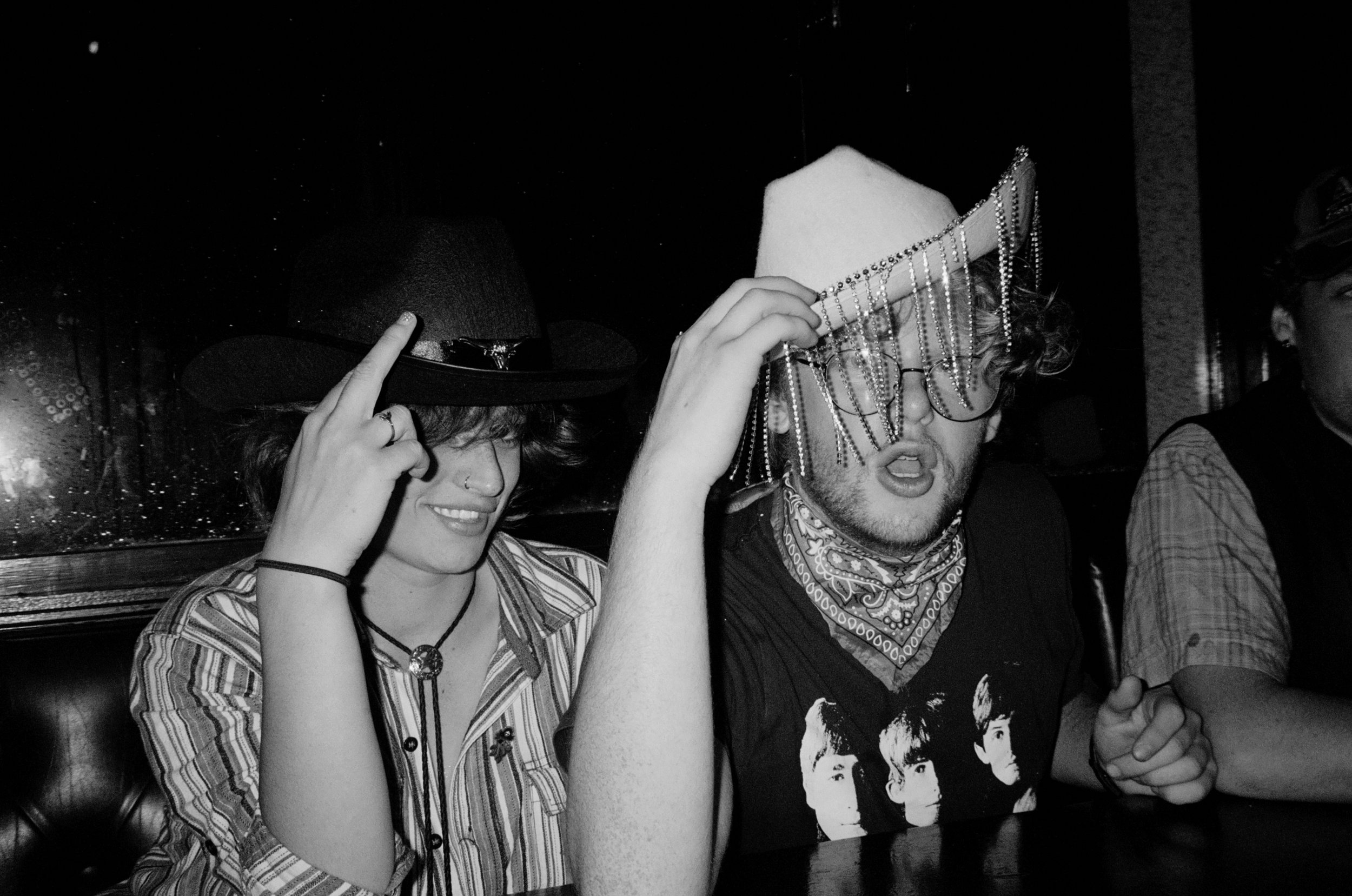 Two young people sitting together, one wearing a wide-brimmed hat and the other holding a beaded mask in front of their face, in a dimly lit setting.