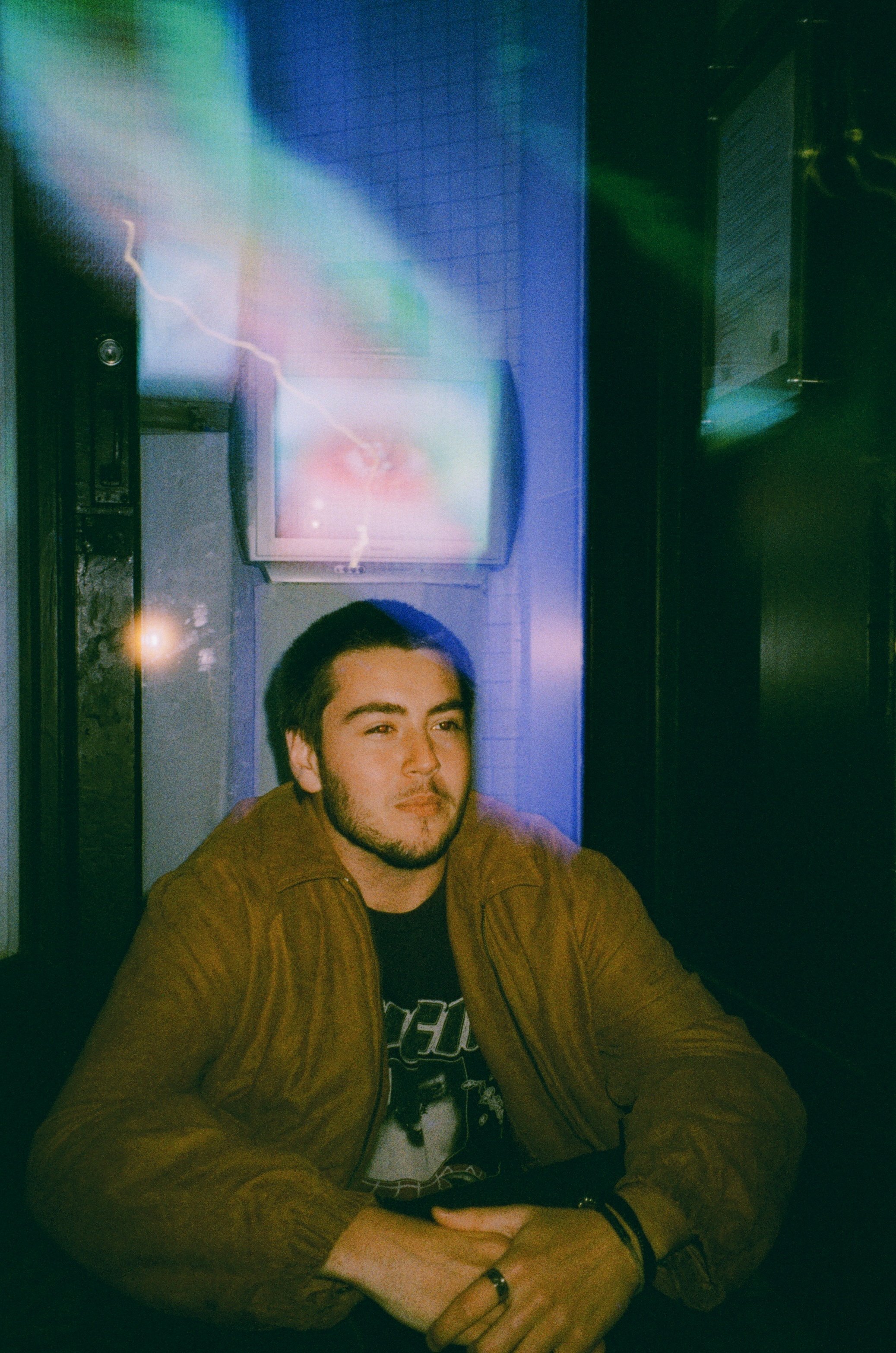 A young man with dark hair, beard, and mustache, sitting at a table with arms crossed, wearing a brown jacket over a black graphic T-shirt, in a dimly lit room with a television screen in the background.