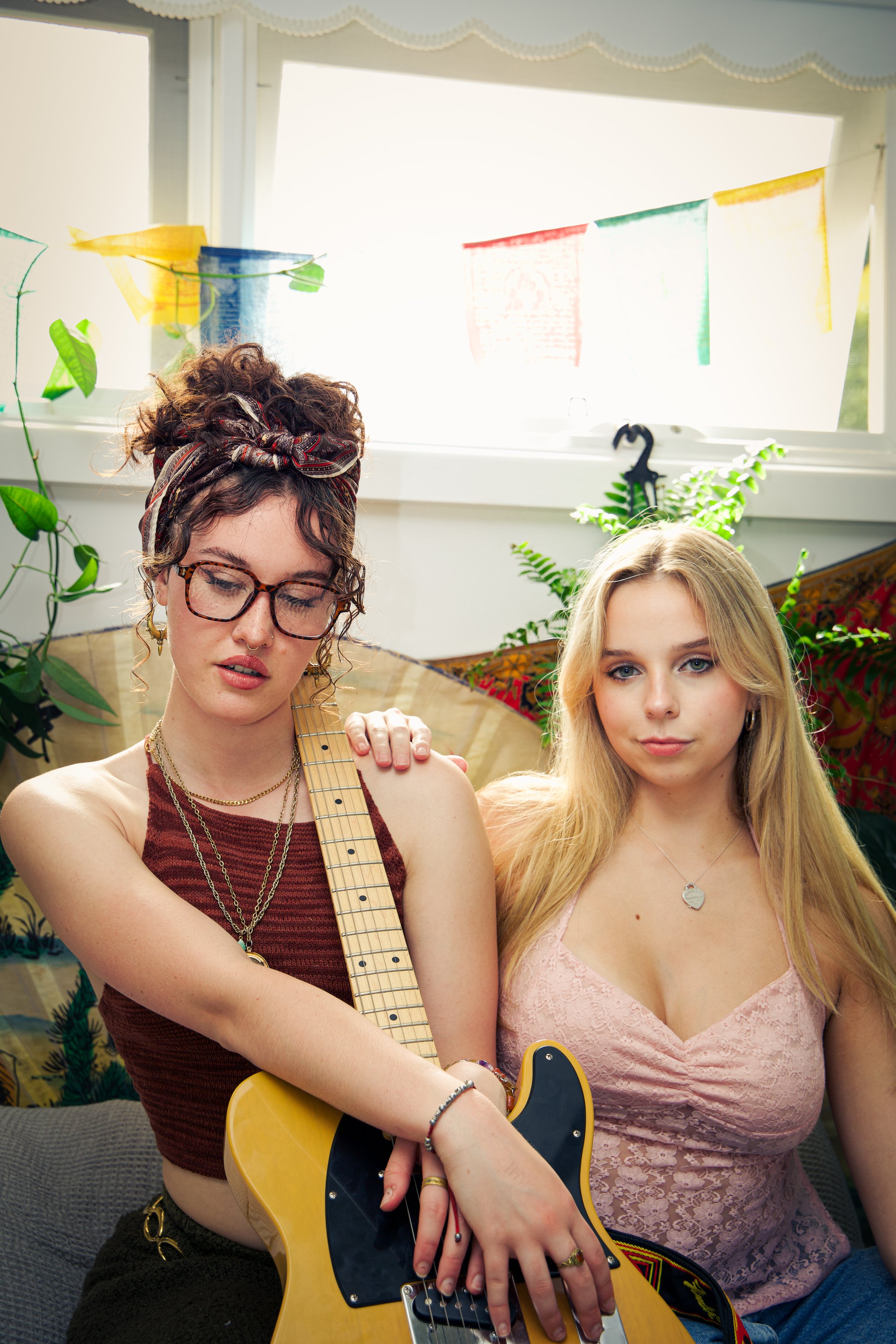 Two young women sitting indoors, one holding a yellow electric guitar, with plants and colorful banners in the background.