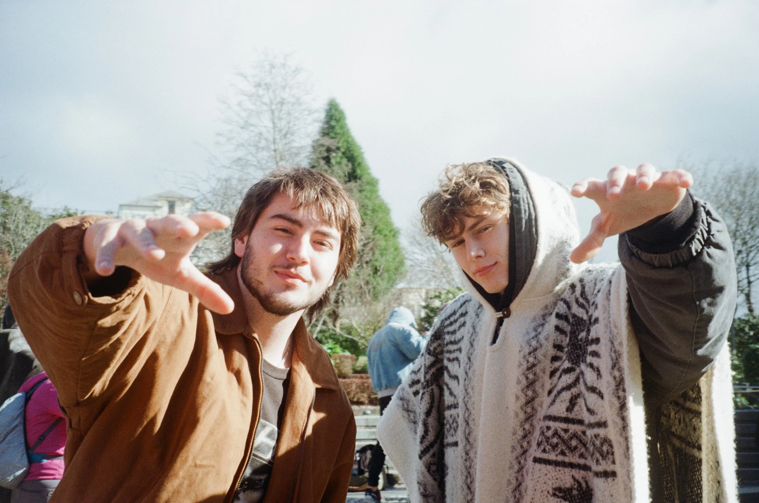 Two young men with curly hair and casual clothing, making hand gestures towards the camera outdoors on a sunny day, with trees and a person in the background.