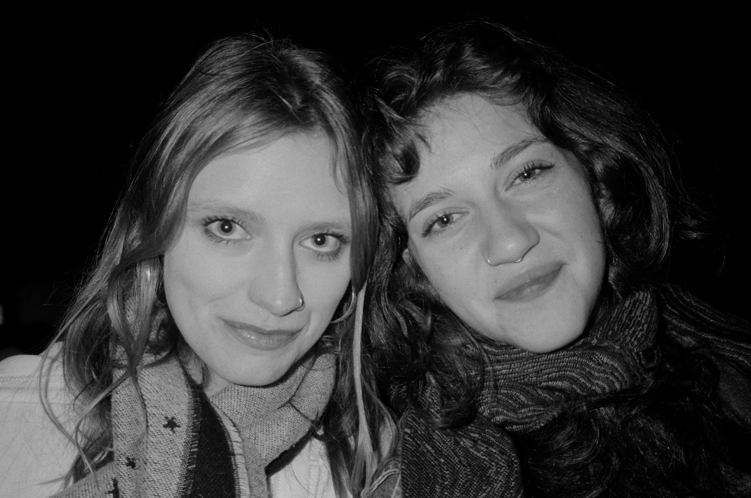 Two women with long hair, one with straight and the other with curly hair, smiling and closely positioned together in a black-and-white photograph.