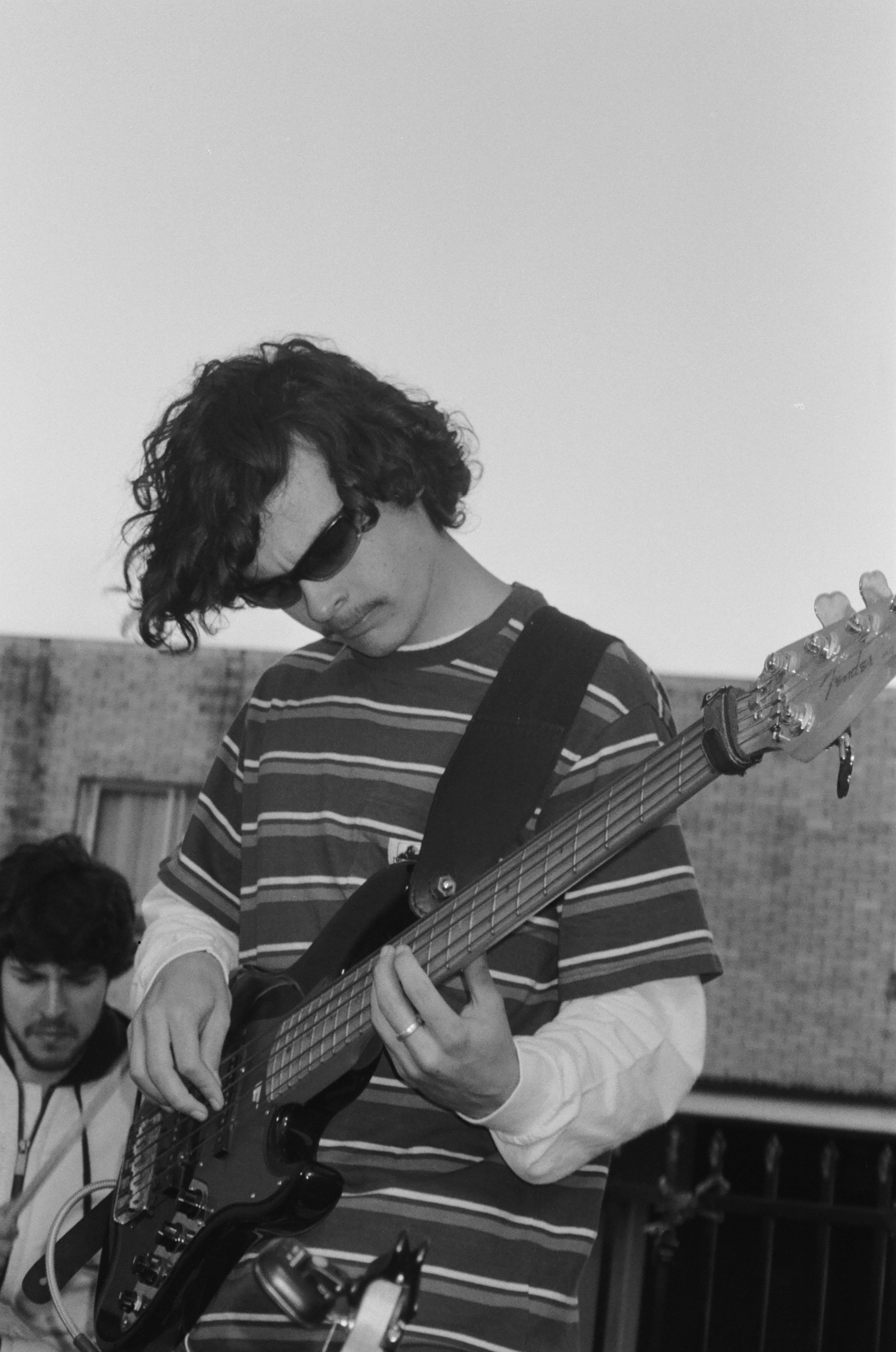 A young person with curly hair wearing sunglasses, a striped shirt, and long sleeve underneath, playing an electric guitar during a music practice or performance, with two other individuals partially visible in the background.