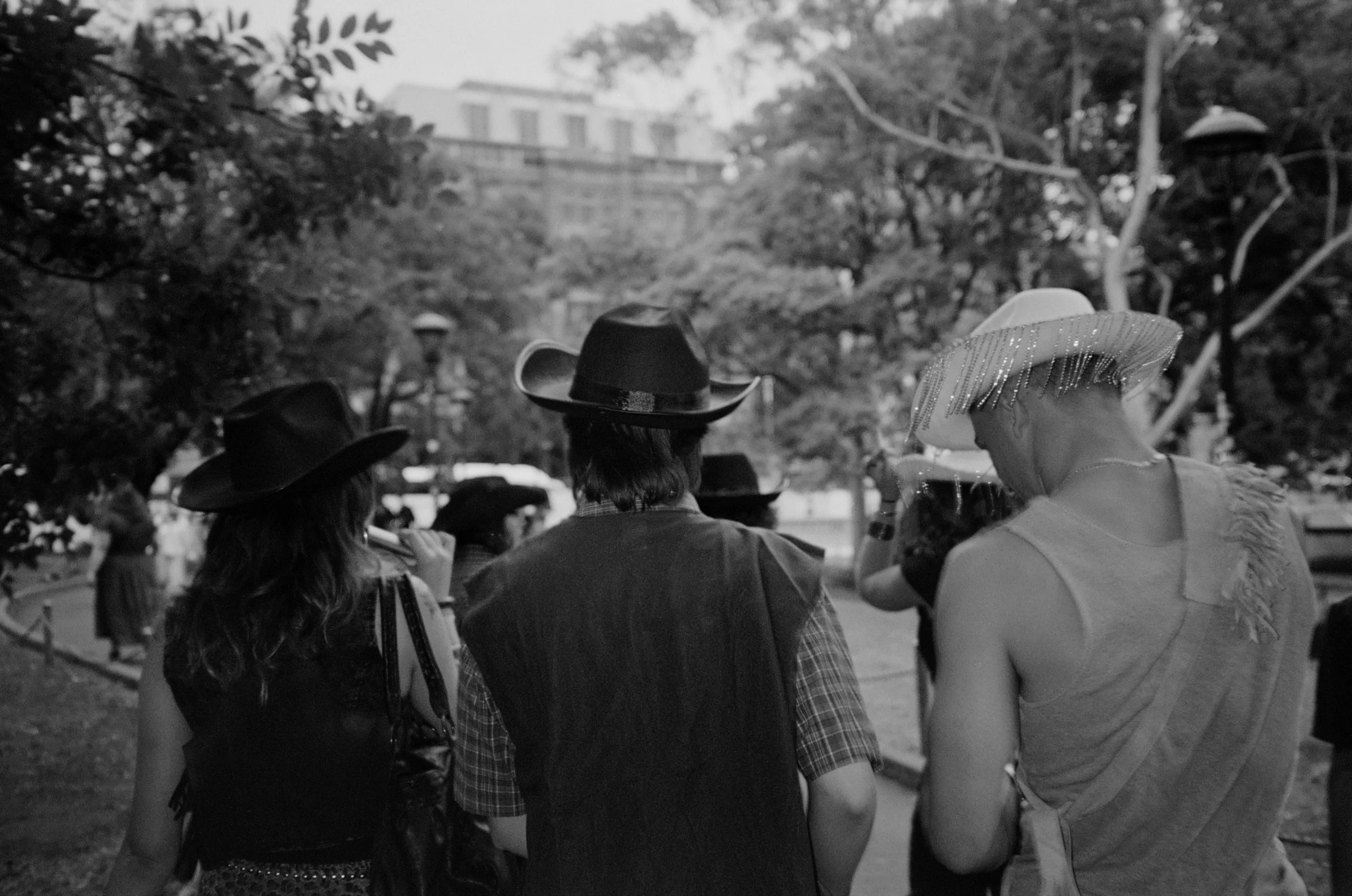 People gathered outdoors, wearing hats, with trees and a building in the background, in a black and white photo.