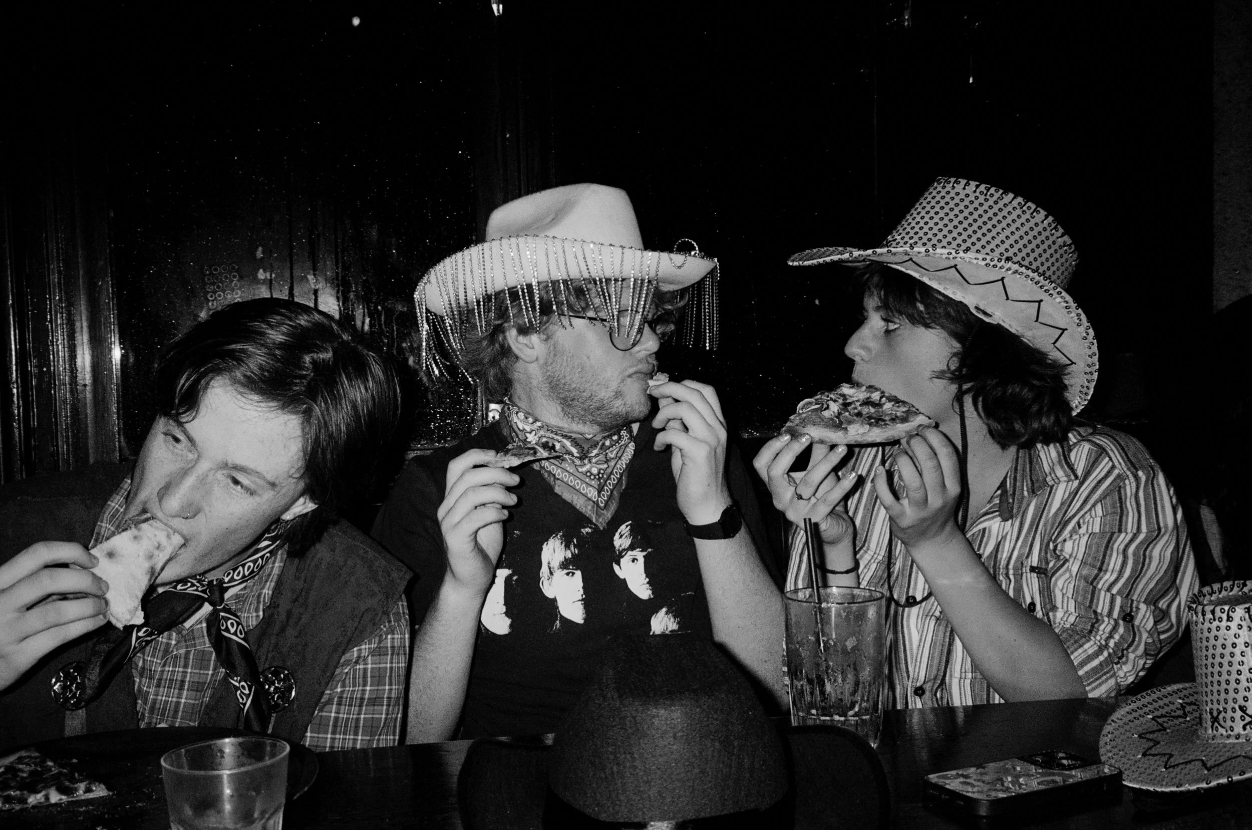 Four people sitting at a table, two women and two men, wearing cowboy hats and bandanas, eating pizza, with drinks on the table.