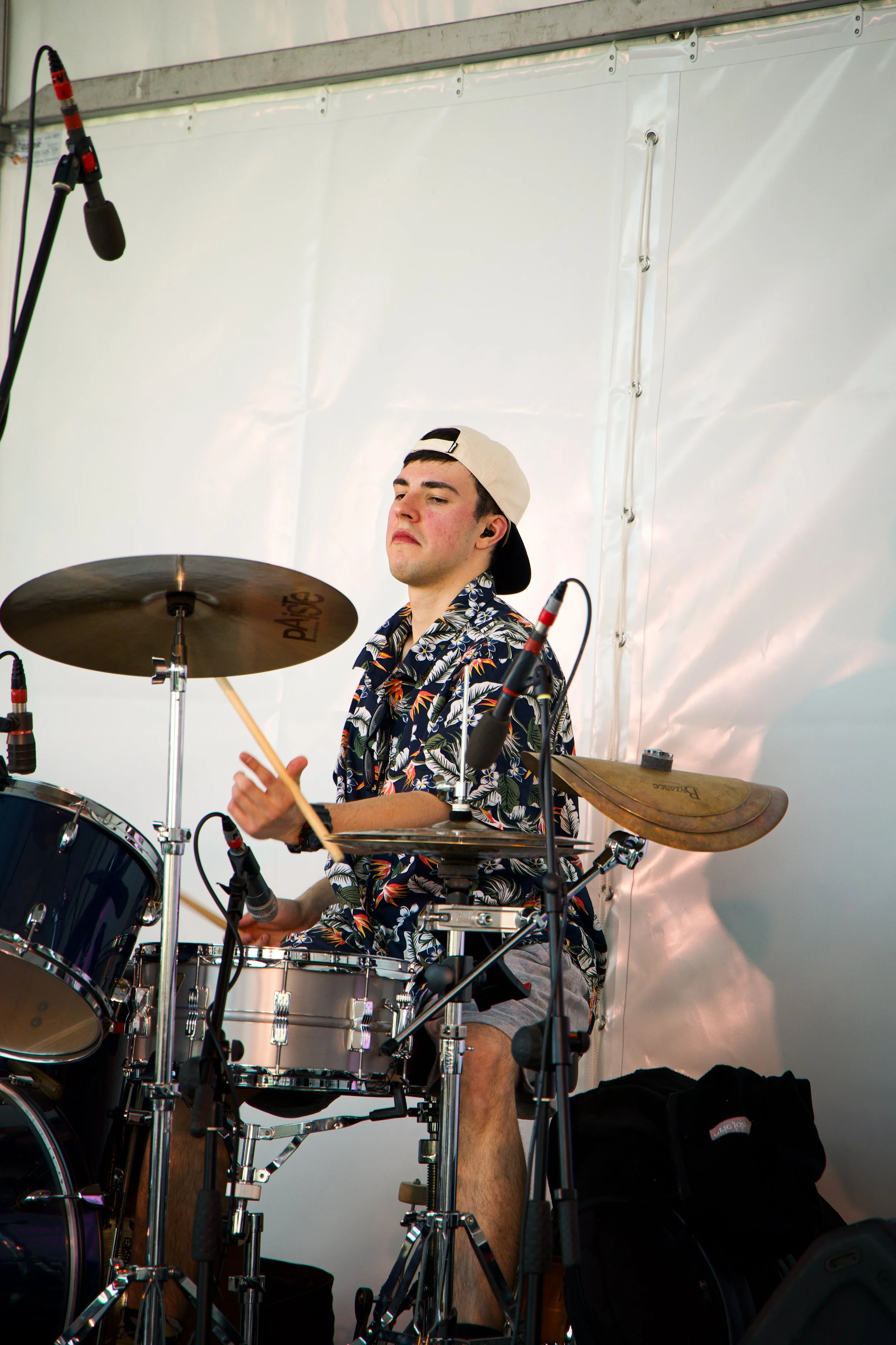 A young man in a tropical patterned shirt and a backwards cap is playing drums on stage, surrounded by microphones and musical equipment, with a white backdrop behind him.