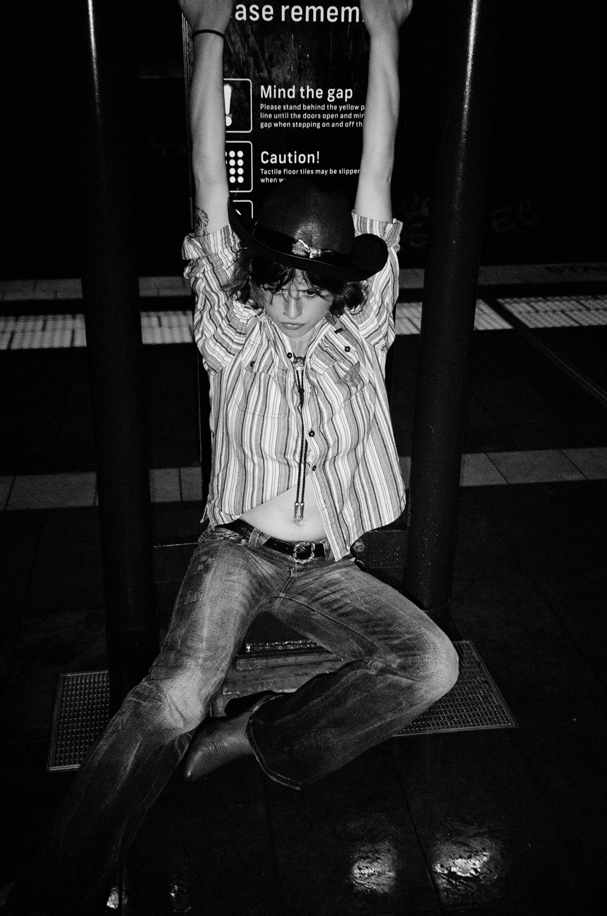 A person wearing a striped shirt, jeans, and a cowboy hat, kneeling on the floor with arms raised, in front of a dark background and an informational sign.