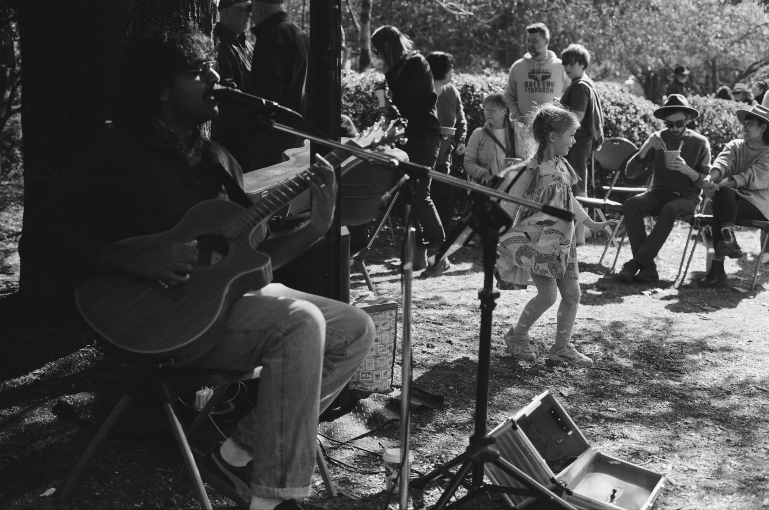 A black and white photo of a group of people outdoors, with a musician playing guitar and singing, and children enjoying the scene, some sitting and chatting, at what appears to be a casual outdoor gathering or festival.