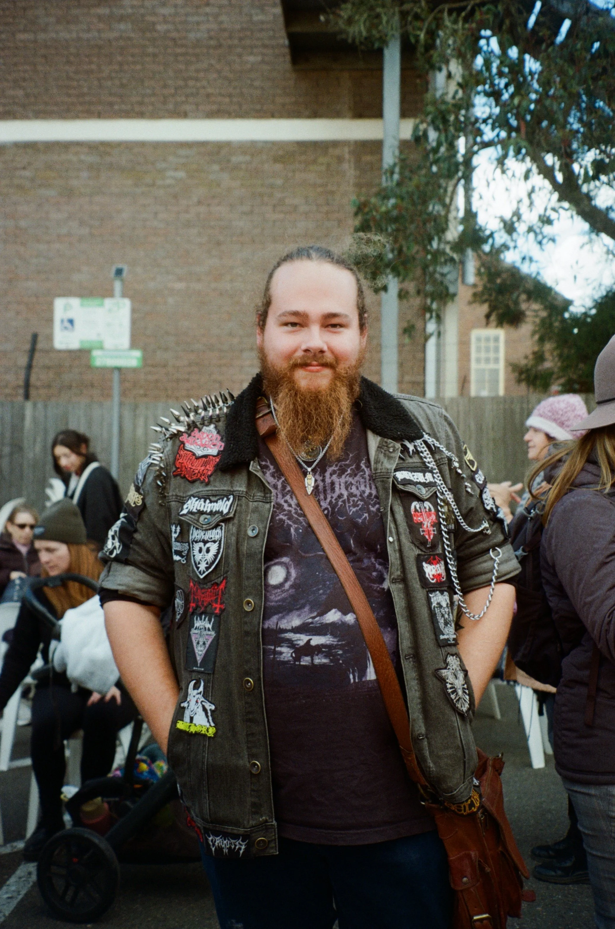 A man with a long beard and long hair tied back, wearing a patch-covered denim jacket and a black T-shirt, stands among people outdoors. The background includes a brick building and trees, and some people are engaged in conversations or activities.