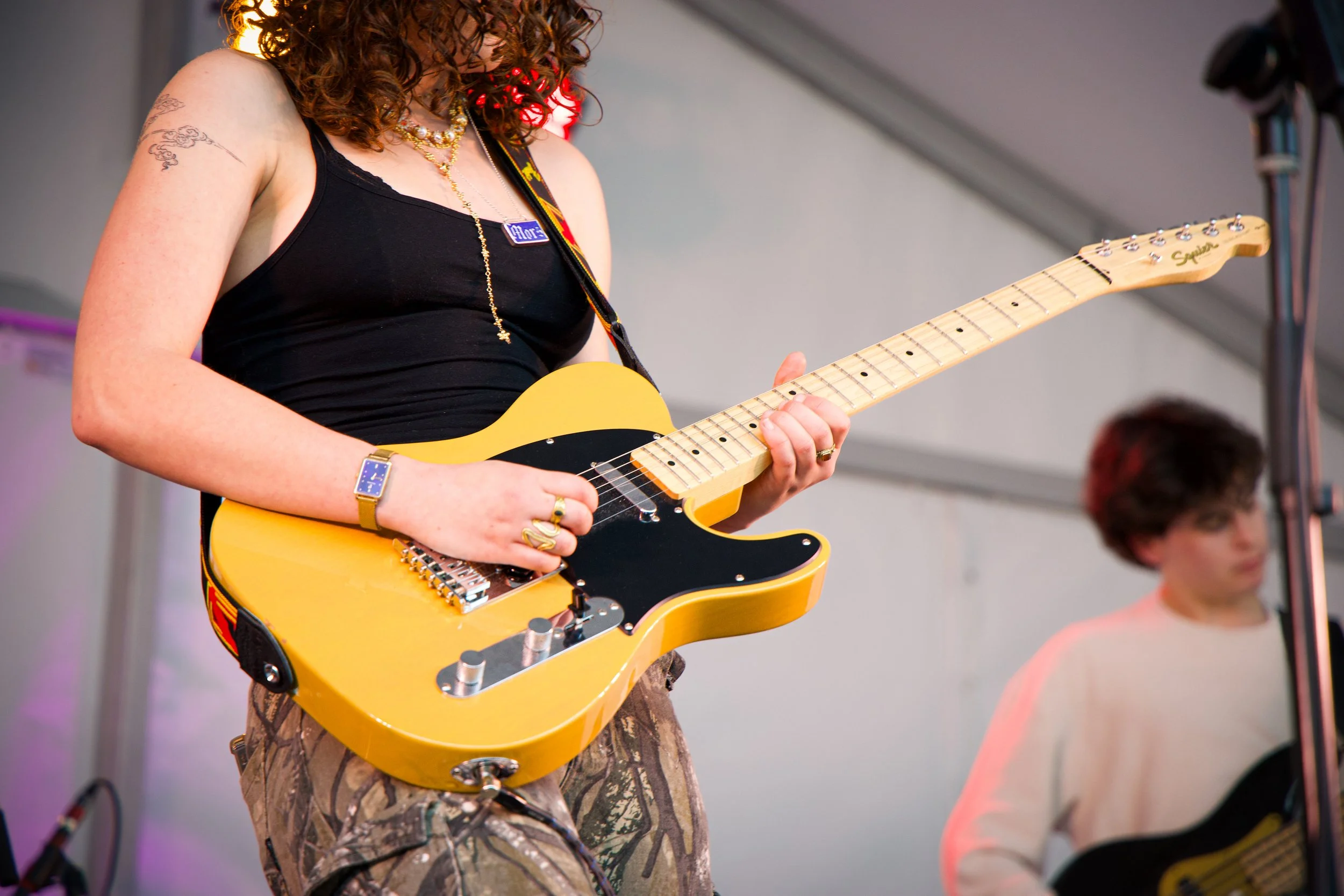 A woman with curly hair wearing a black tank top, a necklace, and jewelry playing an electric guitar on stage. Another person with short hair and a pink shirt sits in the background.