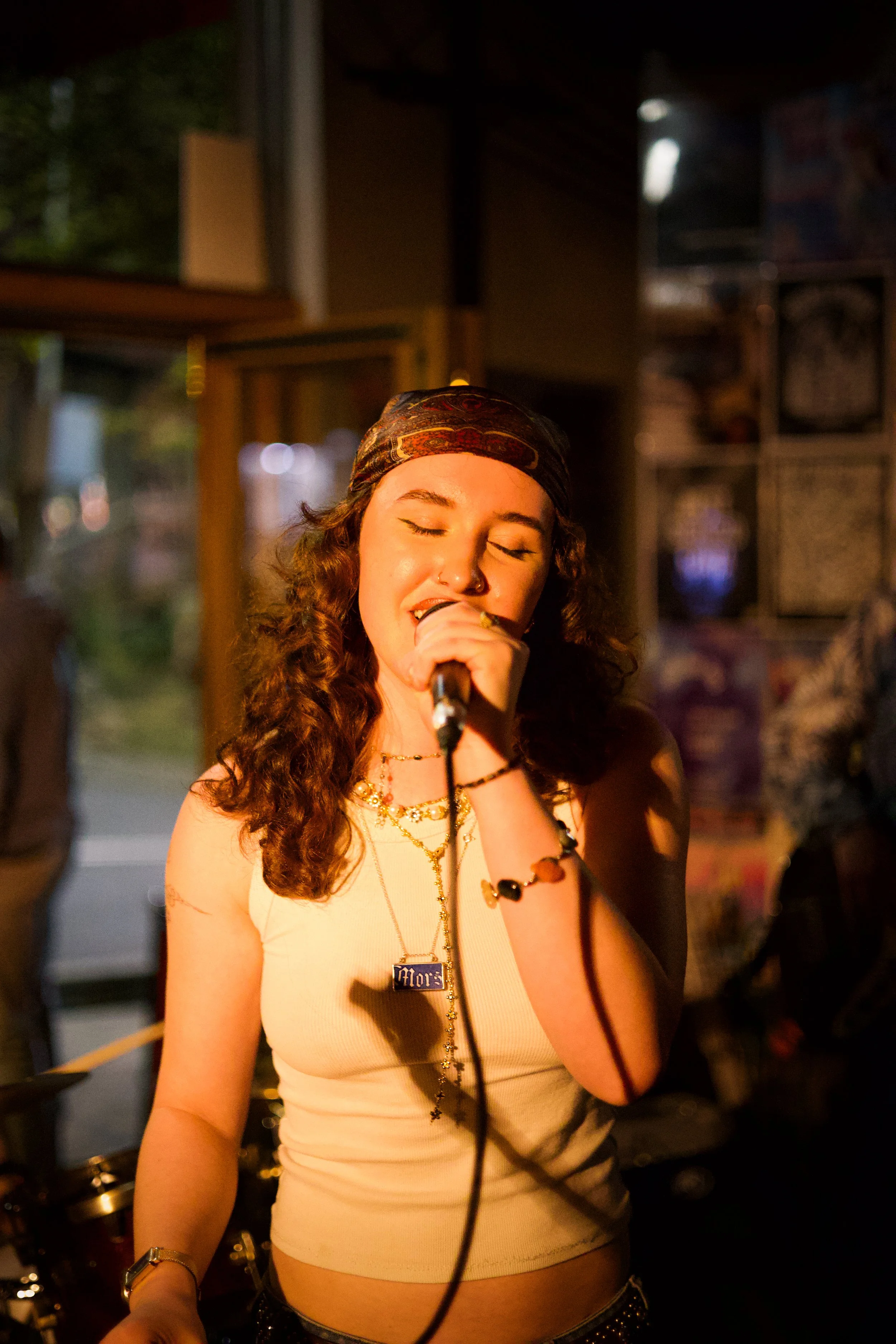 A young woman with curly hair, wearing a bandana and jewelry, singing into a microphone in a dramatically lit indoor setting.