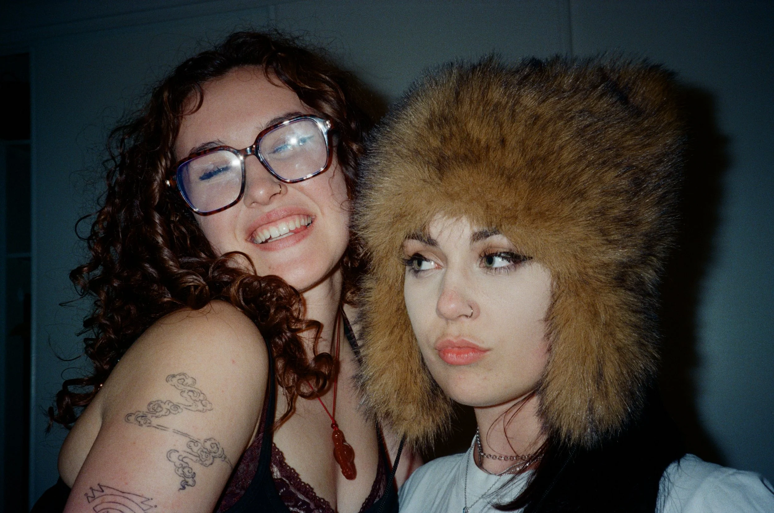 Two women posing closely together at a social event. The woman on the left has curly brown hair, large glasses, a nose ring, tattoos on her arm, and is smiling with her eyes closed. The woman on the right has straight, dark hair under a large fur hat