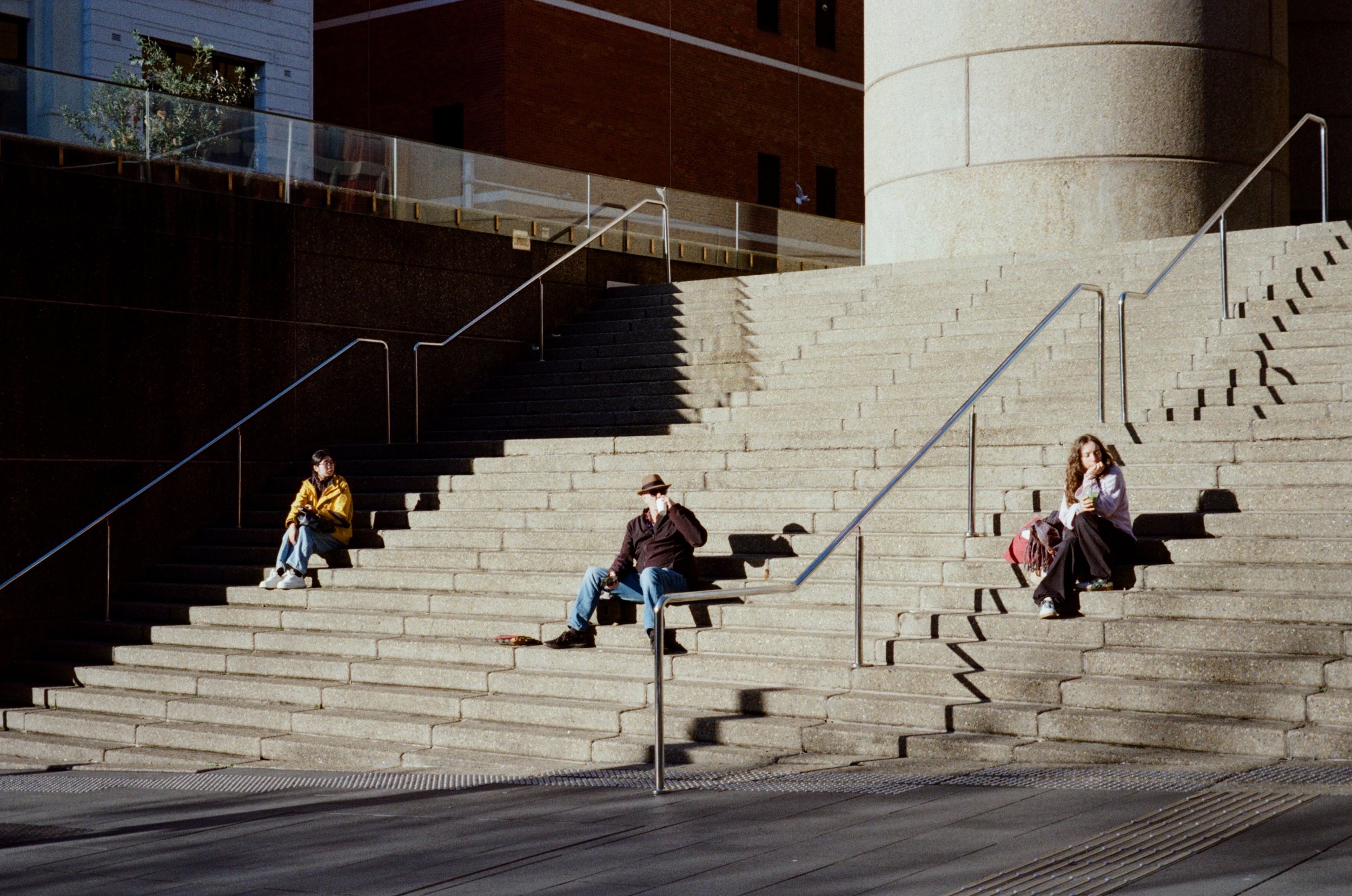 People sitting on wide outdoor stone stairs near a building, with metal handrails and shadows cast by the sunlight.
