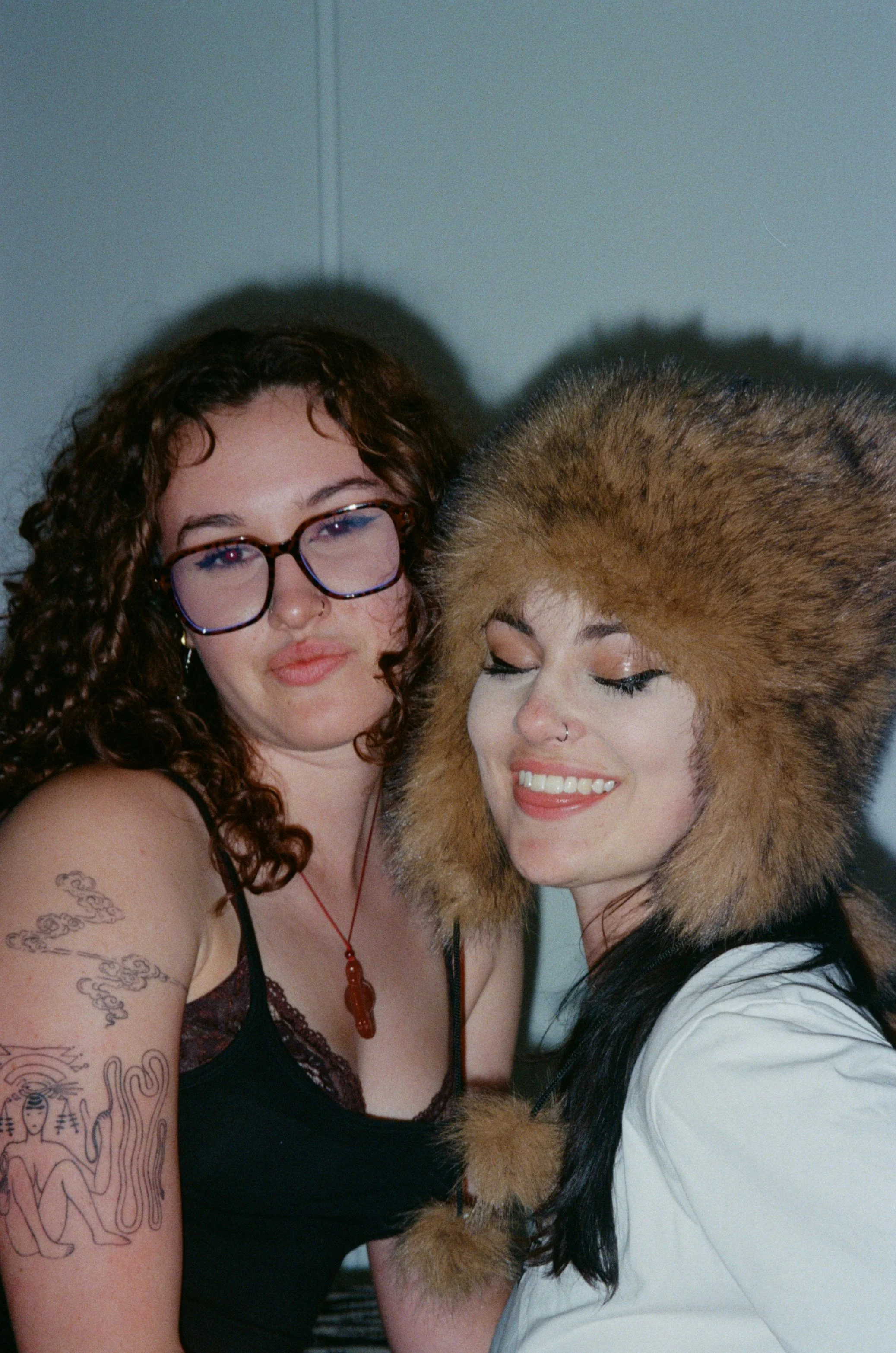 Two women close together, one wearing glasses and a black top with tattoos on her arm, the other wearing a furry hood and white top, both smiling with their eyes closed.