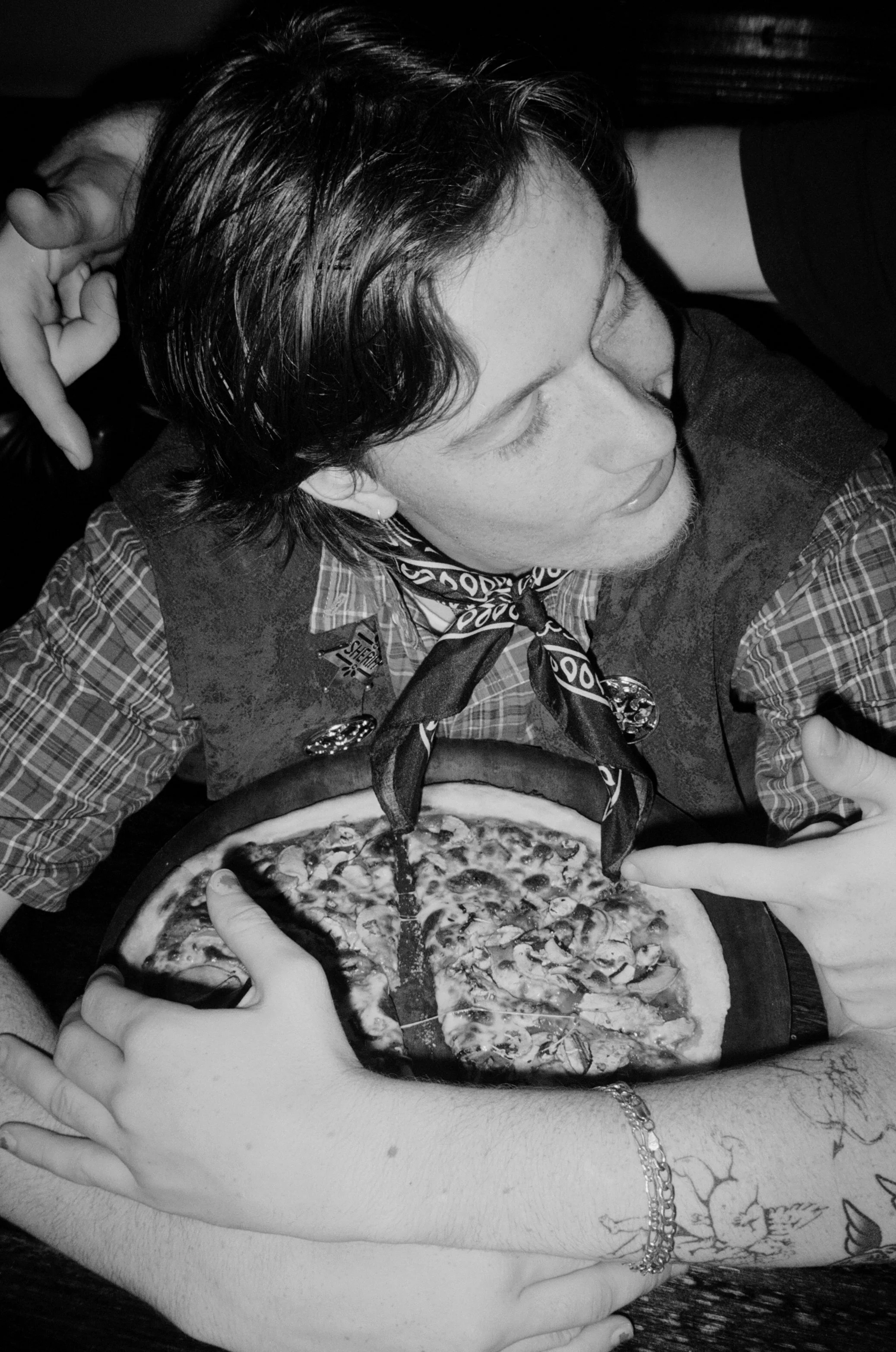 A person with tattooed arms and wavy dark hair is sitting at a table, looking down at a pizza in front of them. Another person’s hand is reaching in to hold the pizza.