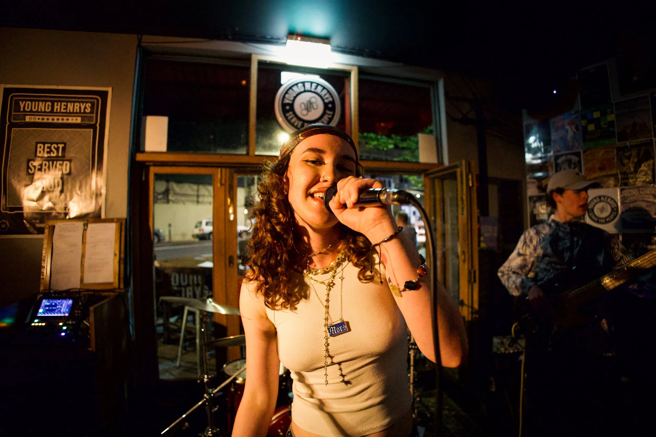A young woman singing into a microphone inside a dimly lit venue with posters on the wall and a man playing guitar in the background.