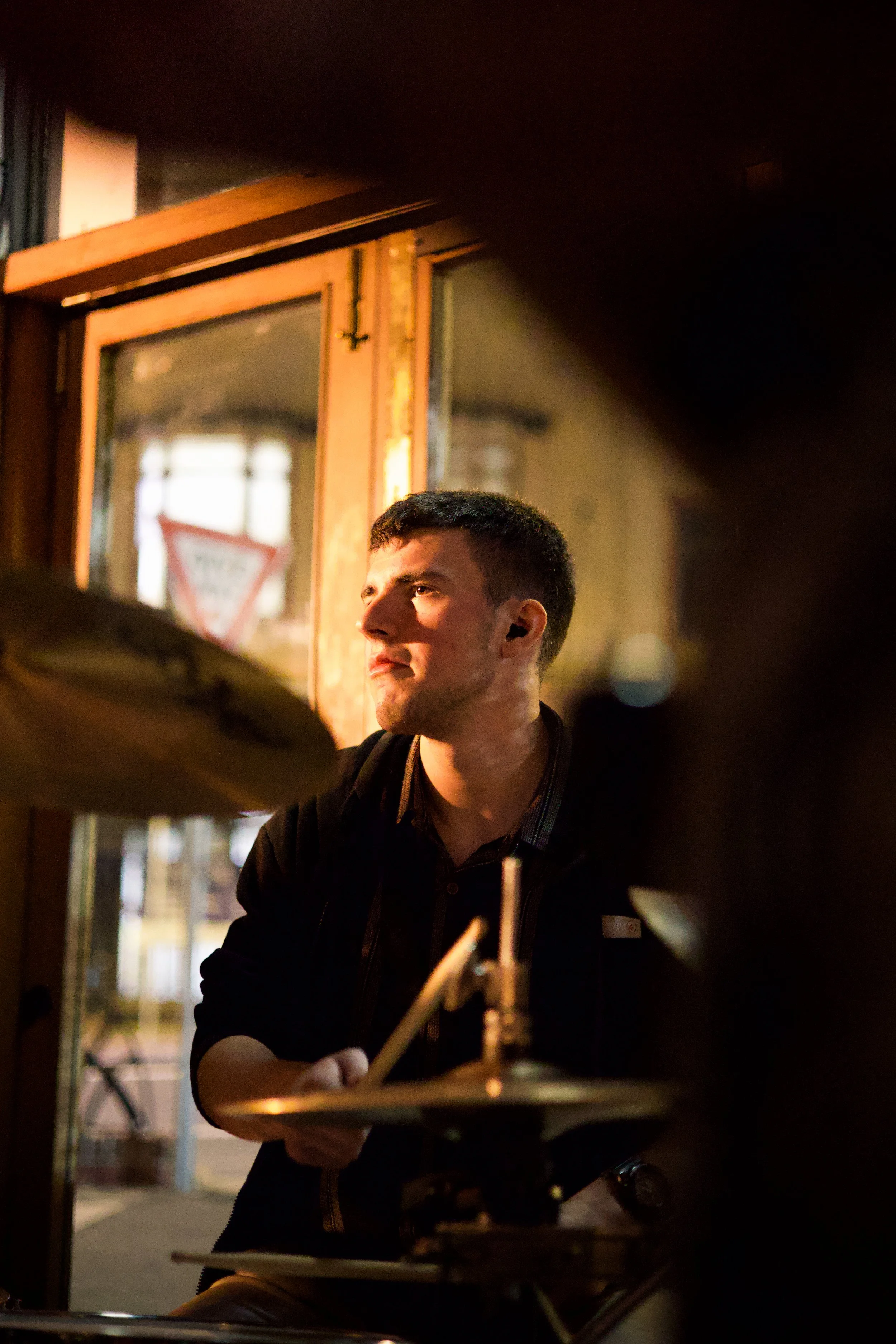 A young man with short dark hair and a black jacket sitting in a dimly lit restaurant or bar, with a large mirror and wooden frame in the background, and partially visible drums or musical equipment in the foreground.
