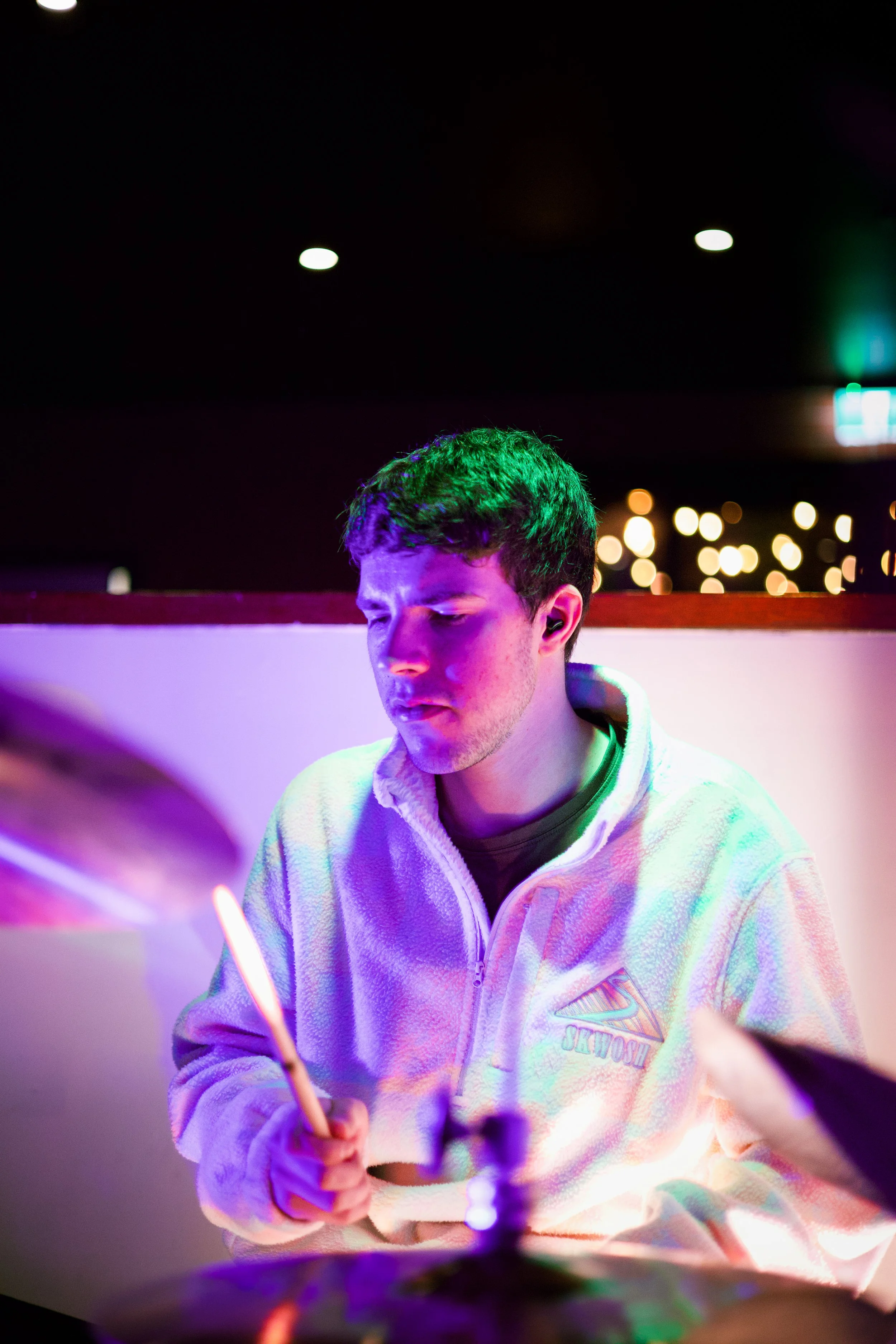A young man with dark hair, wearing a white fleece jacket with a logo, is in a dark room with colorful lights, holding a lit sparkler in his hand.
