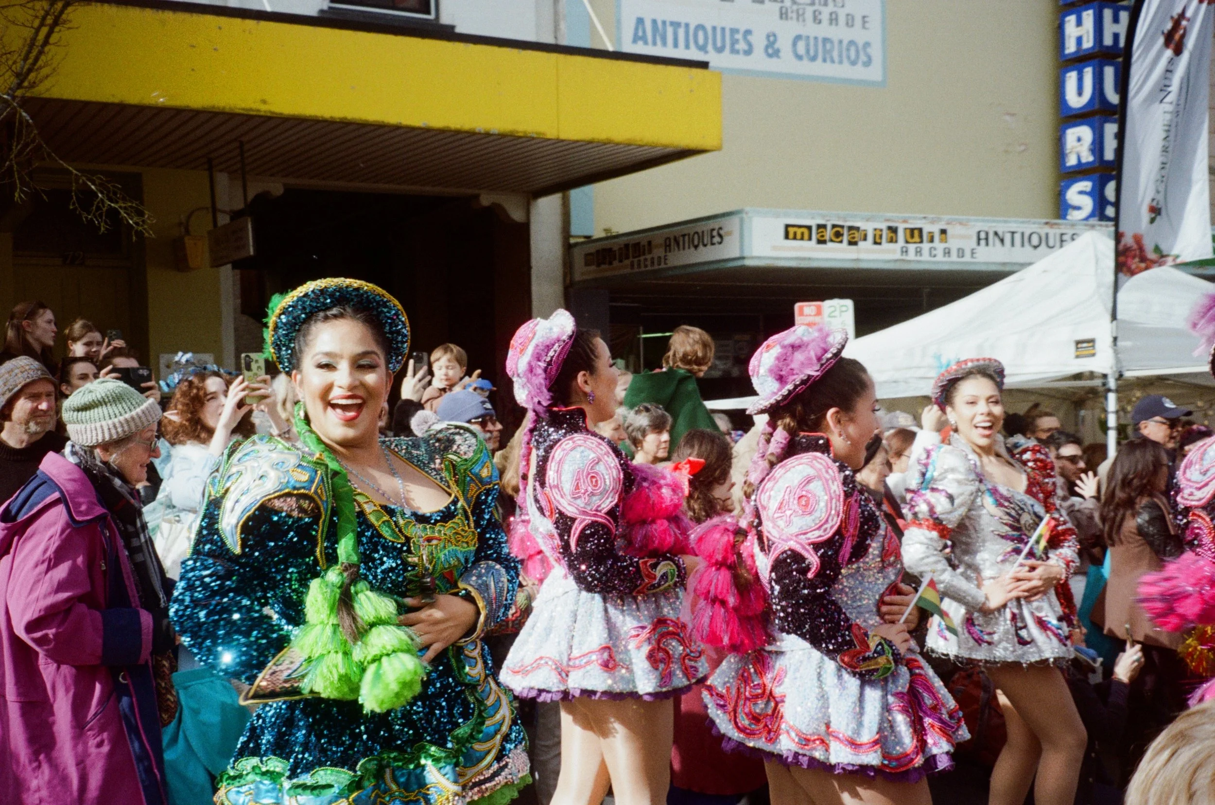 Crowd of people participating in a parade, some wearing traditional colorful costumes with hats and embellishments, in front of a shop with signs reading "ANTIQUES & CURIOS" and "macarthur ARCADE".