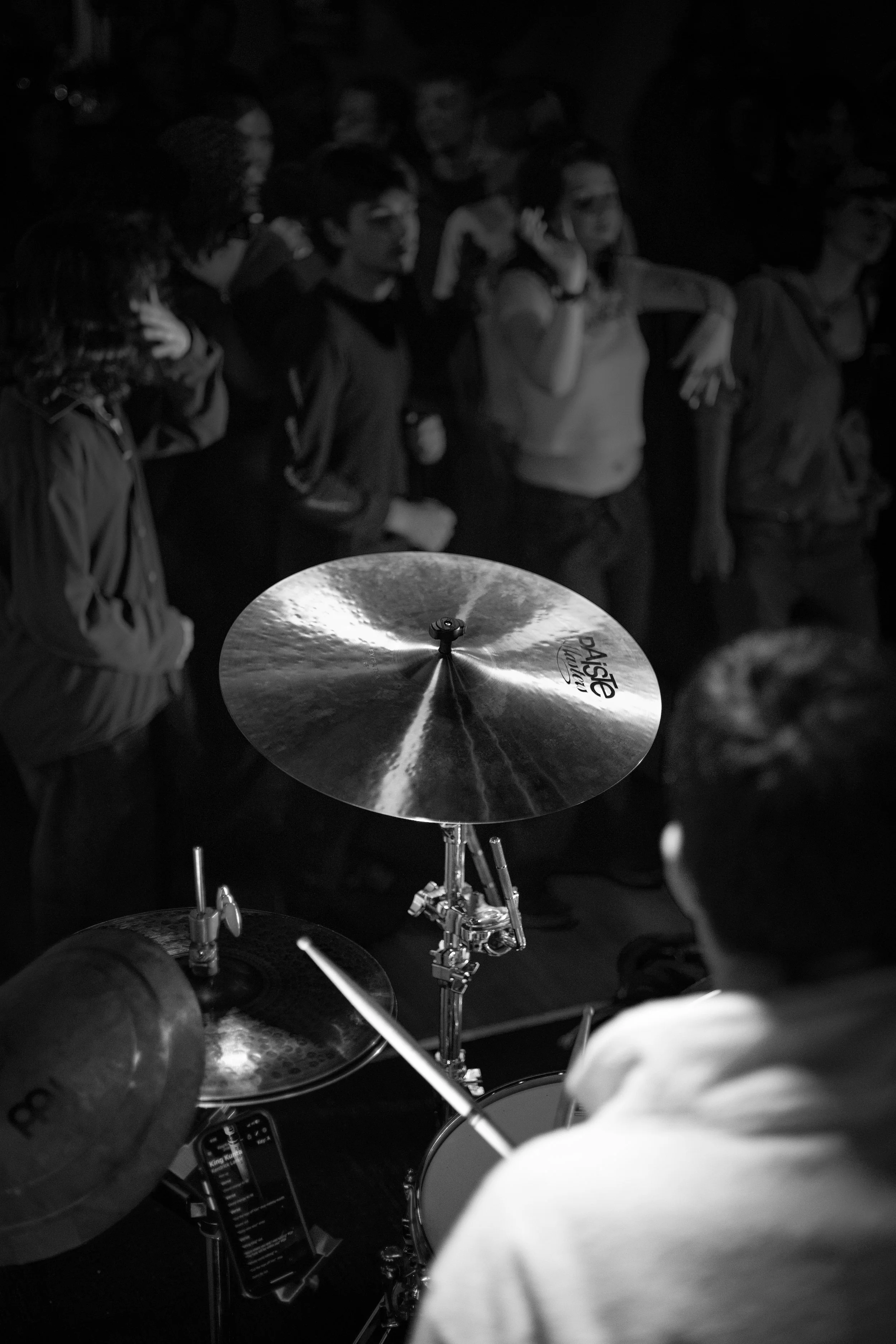 A group of people gathered around a drum set, with a focus on a cymbal labeled Paiste, in a dark setting.