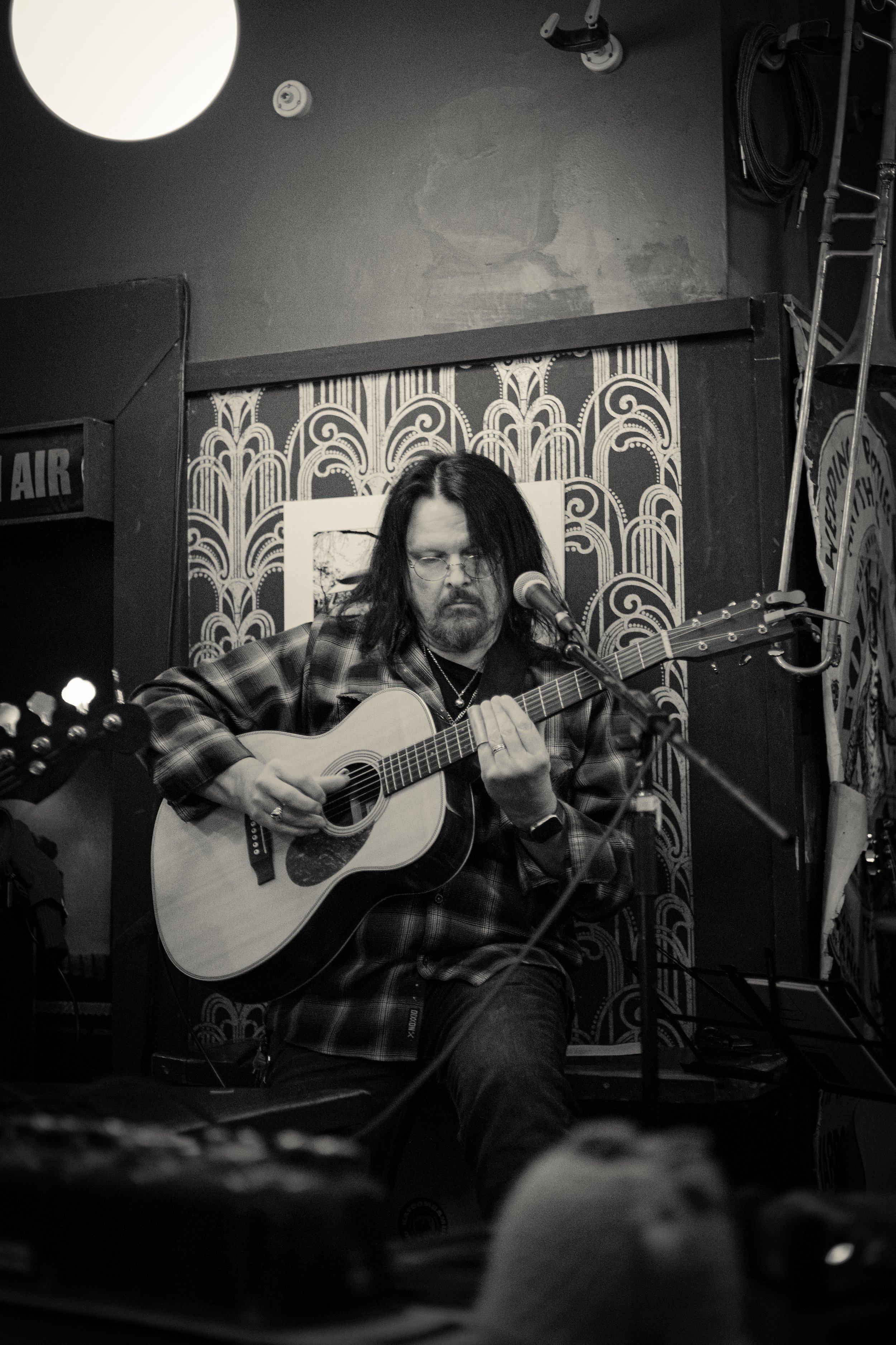 A man with long dark hair and glasses playing an acoustic guitar during a performance in a dimly lit venue with decorative wallpaper.