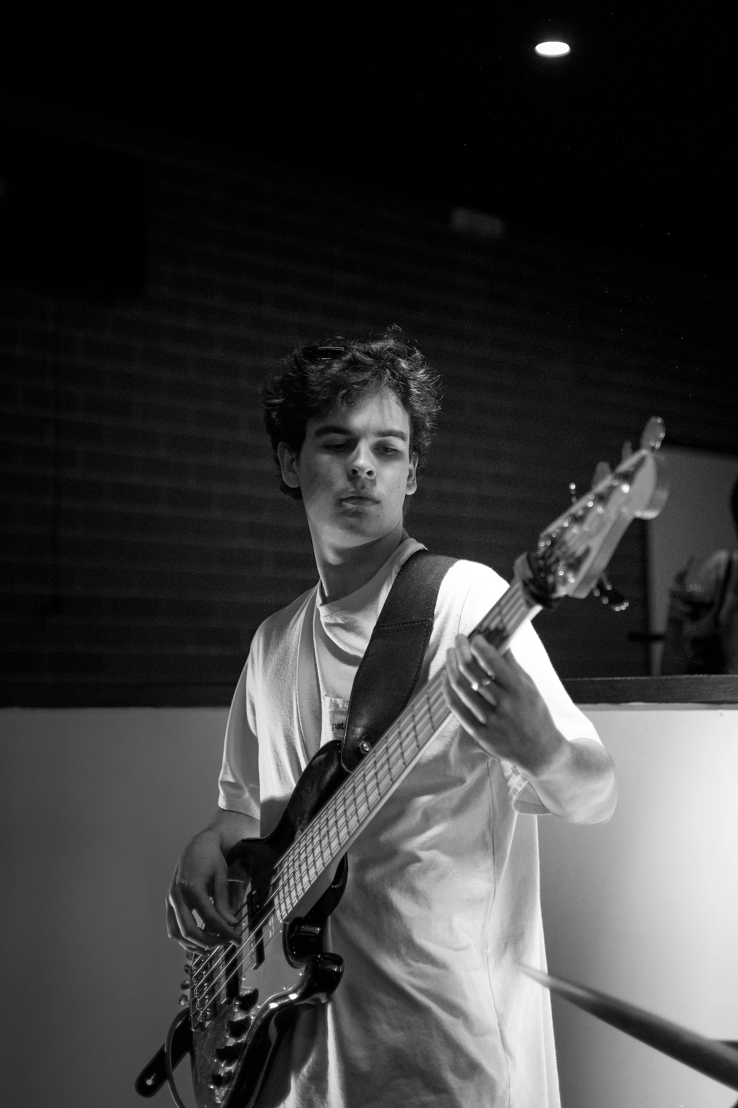 Young man playing an electric bass guitar in a recording studio, wearing a light-colored t-shirt, with a focused expression, and dark background with ceiling lights.