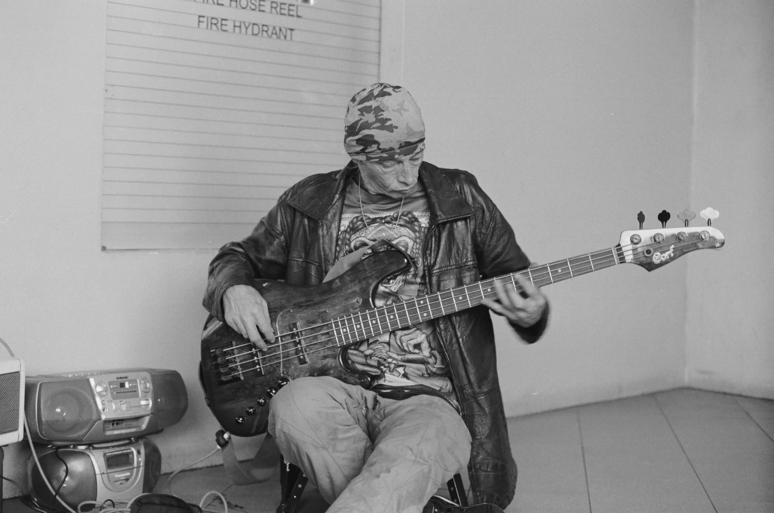 A person with a camouflage bandana, wearing a leather jacket and graphic t-shirt, playing an electric bass guitar while seated on a chair. There is a portable stereo on the floor and a blank lined paper on the wall behind them.
