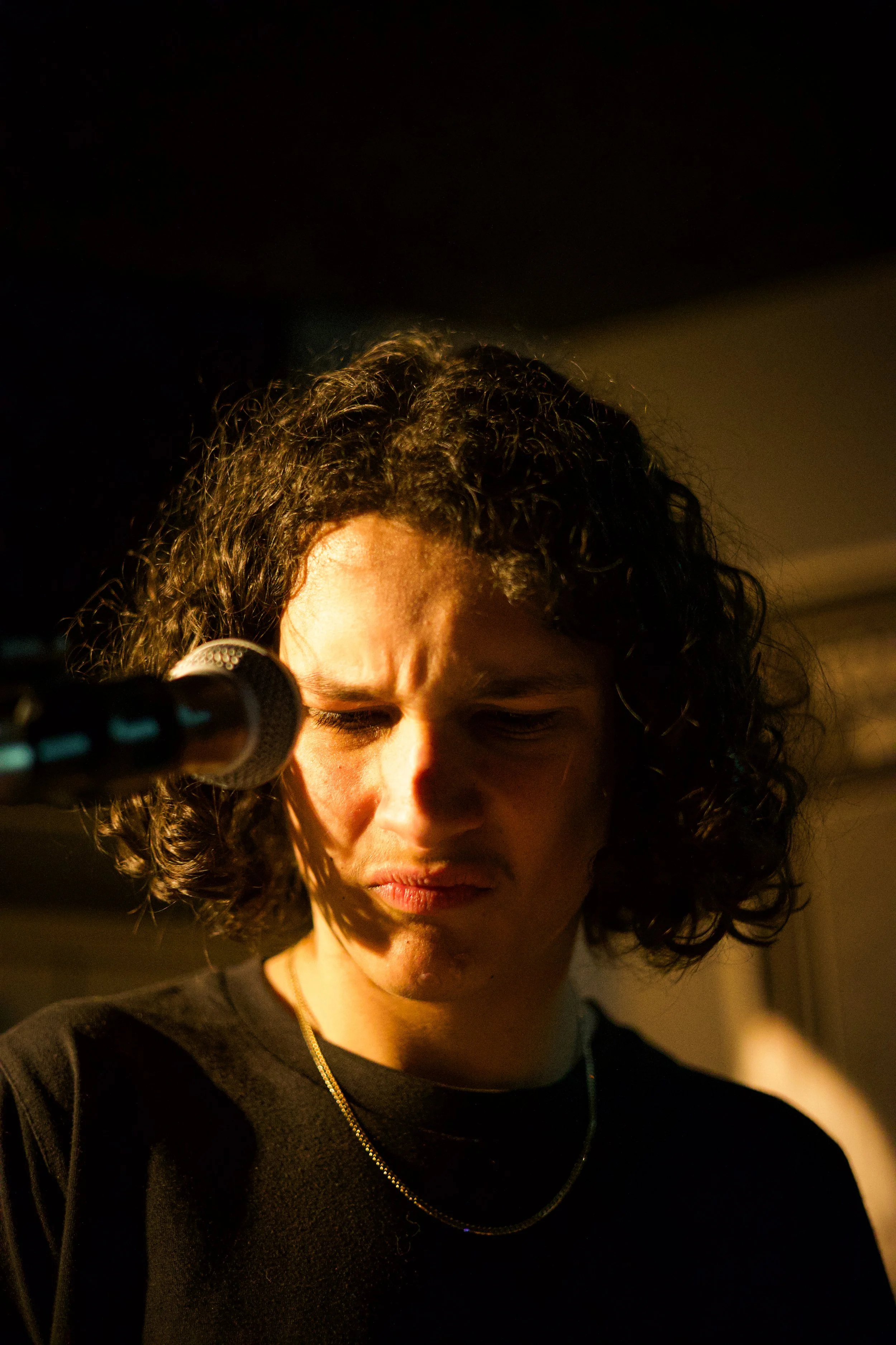 A man with curly hair is standing in front of a microphone with her eyes closed, in a dimly lit room.