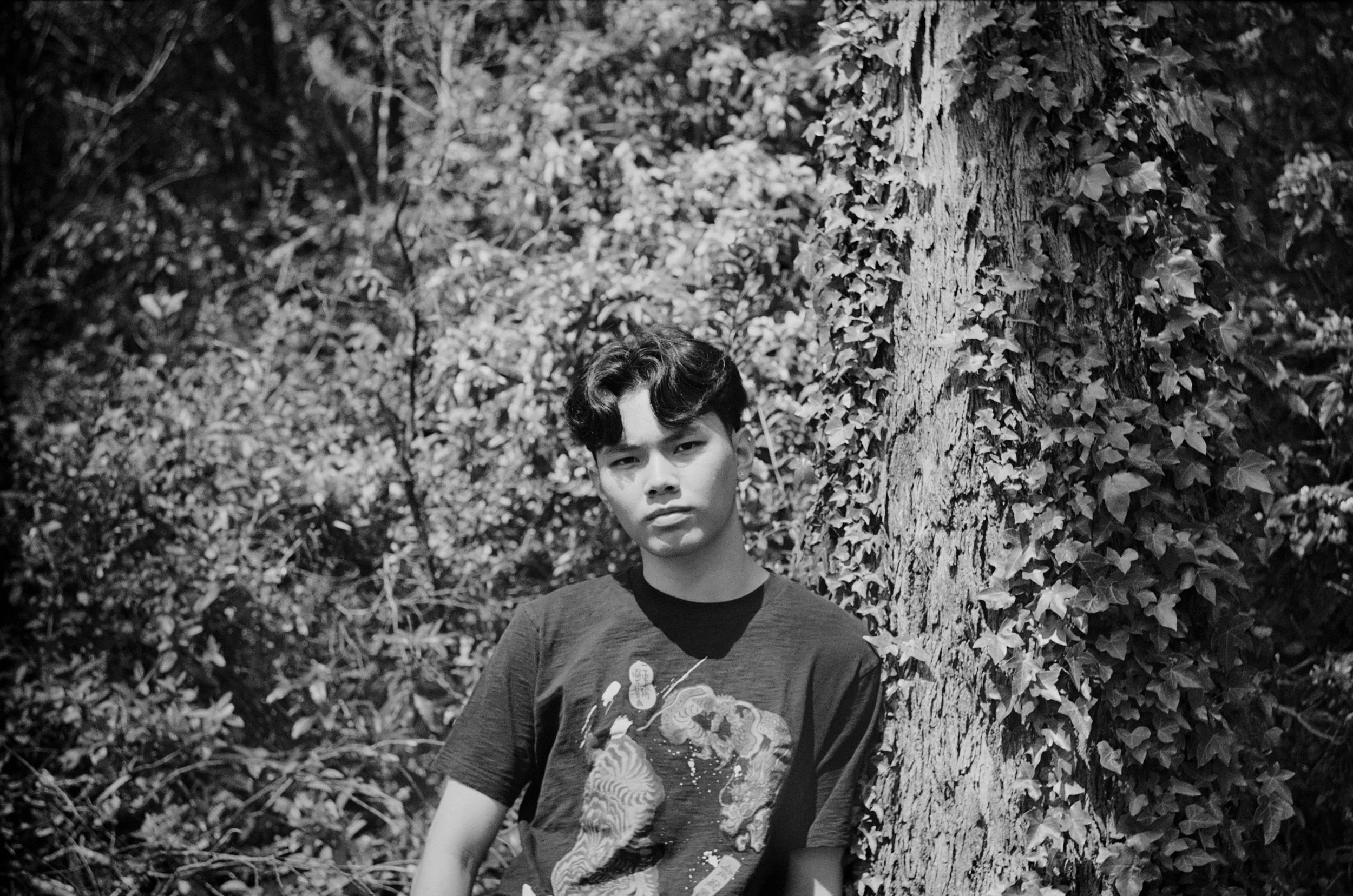 A young man with curly hair standing outdoors next to a large tree covered in ivy, with dense foliage in the background, in black and white.