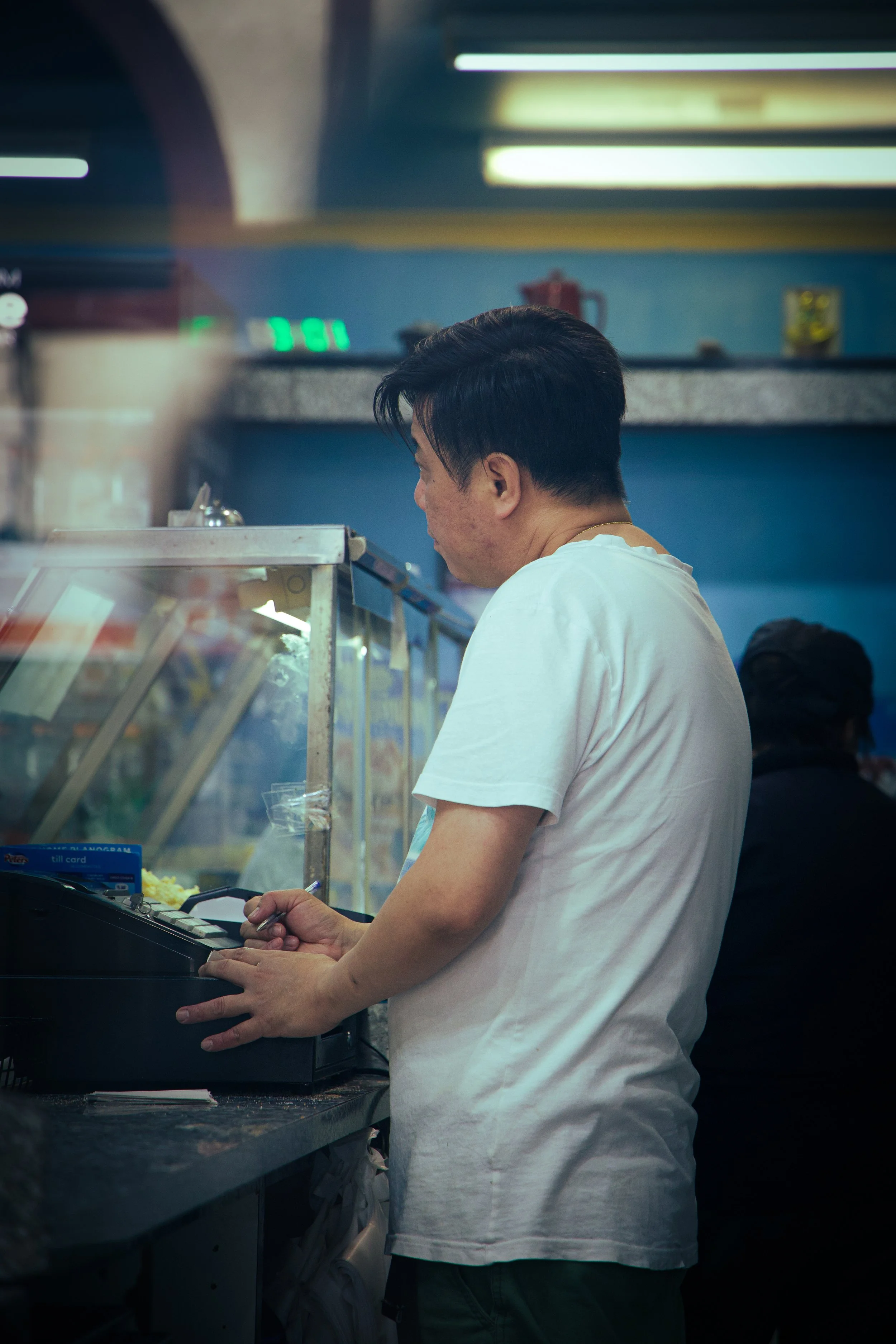 A man wearing a white t-shirt is standing at a counter, operating a cash register, with a glass display case nearby. There are some items inside the display case, and the background has a counter with a fire extinguisher and some green-lit electronic devices.