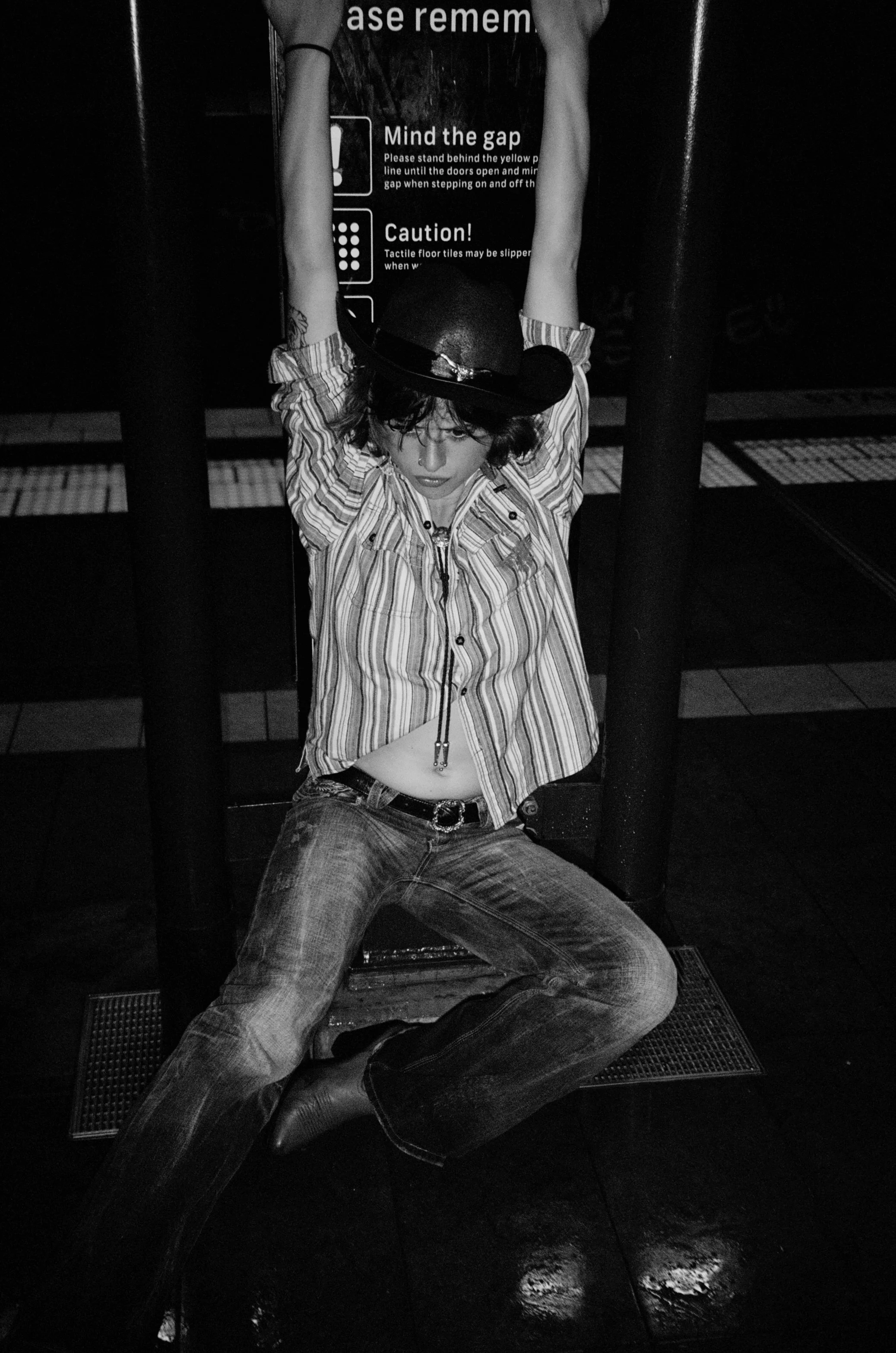 A person with curly hair wearing a cowboy hat, striped shirt, bolo tie, and jeans is sitting on the floor with legs spread apart and hands raised above their head, leaning against a black railing. Behind them is a sign with text visible.