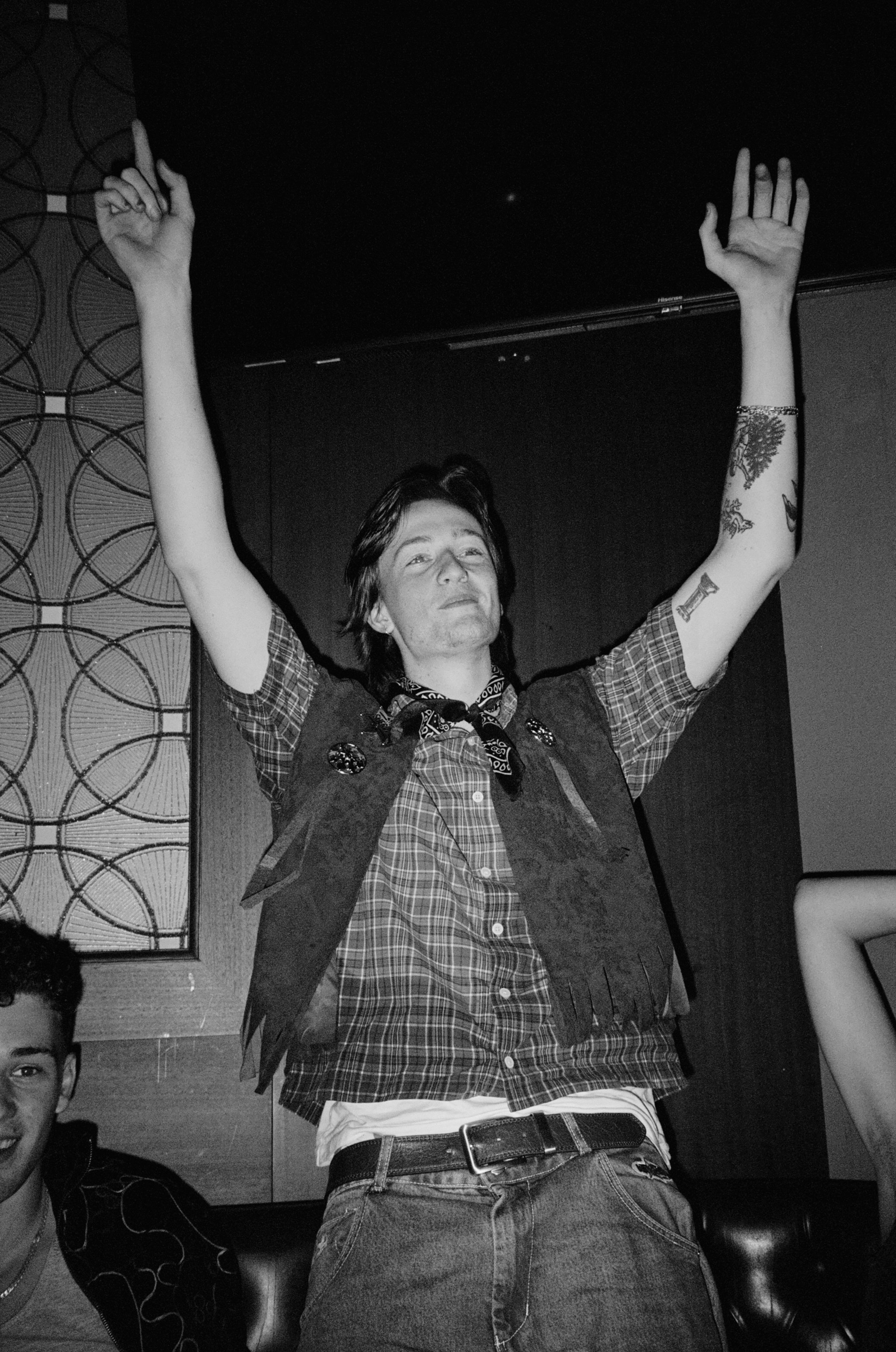 A black and white photo of a man with medium-length dark hair, raising his arms in the air, making a peace sign with his right hand. He is wearing a plaid shirt, a vest, and a neckerchief. There is a tattoo on his left arm. Part of another person's f