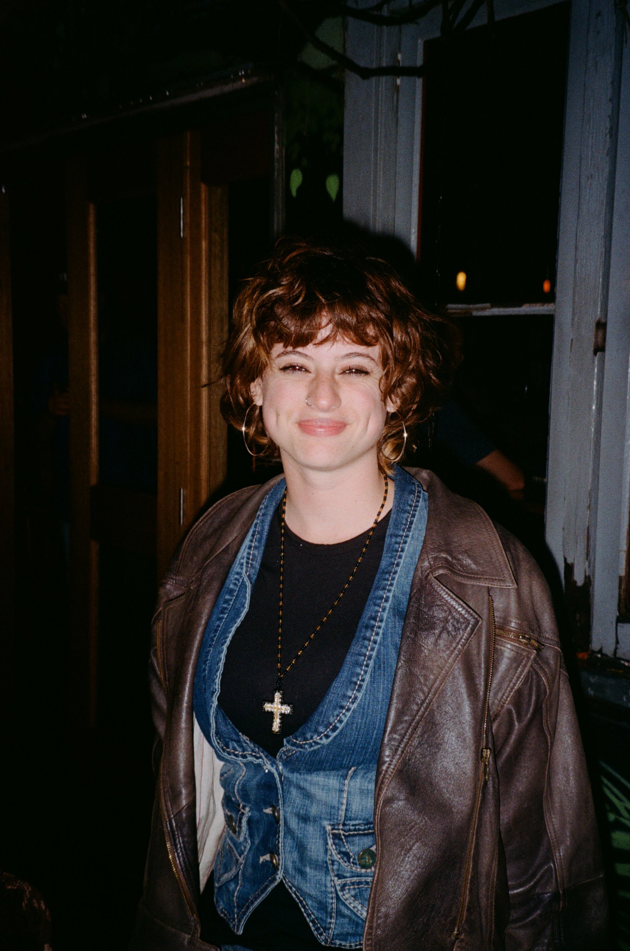A woman with curly brown hair smiling at night, wearing a black shirt, denim vest, and brown leather jacket, with a cross necklace in an outdoor bar setting.