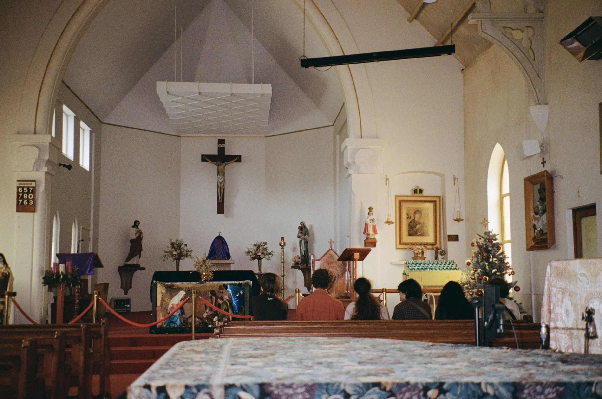 Interior of a church decorated for Christmas with a altar, nativity scene, and Christmas trees, with a young youthful seated in the frontmost pews.